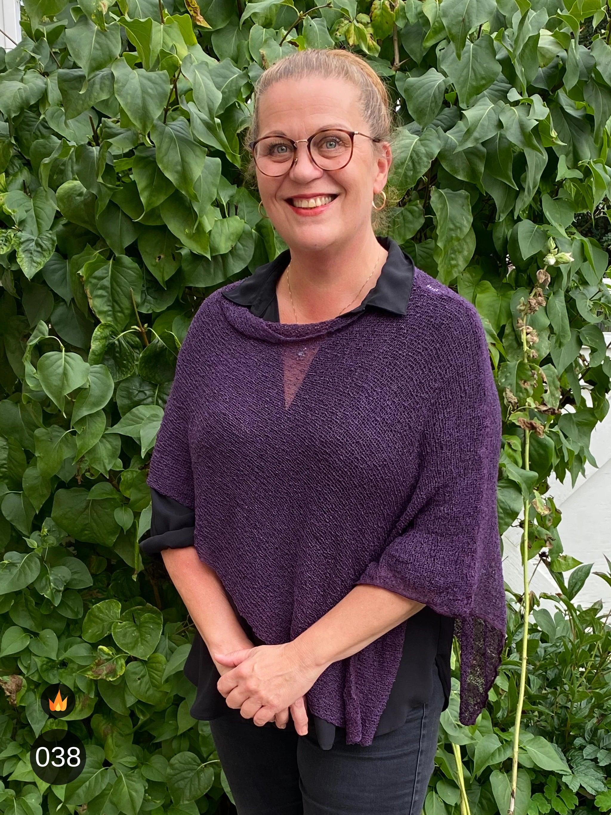 Kvinna bär Ella Cape i färg 038 Aubergine, en varm färg i djup aubergine, finstickad cape som draperas mjukt över axlarna, fotad framför en grön murgrönshäck. Ulla Jacobsson Kläder.