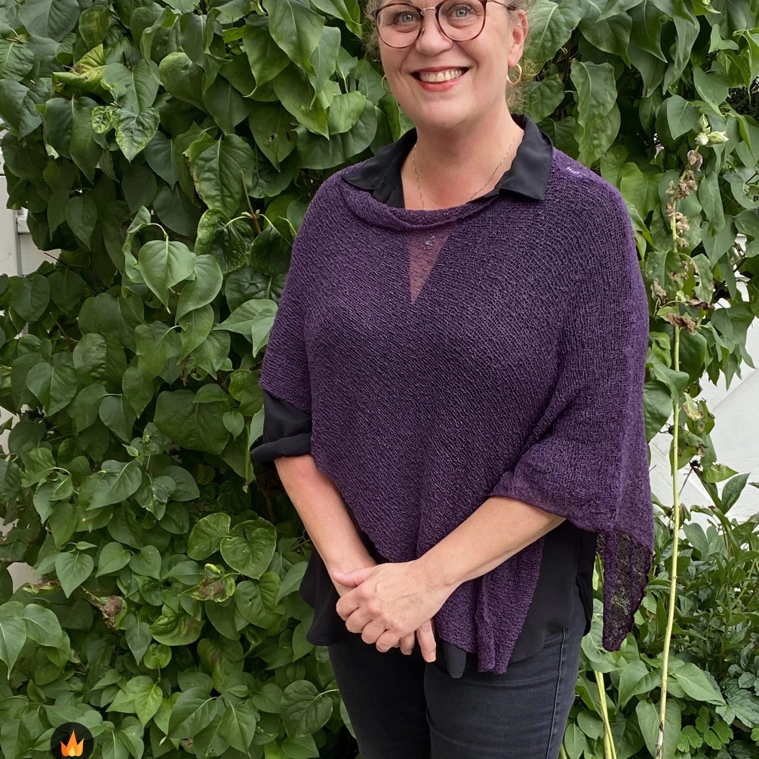 Kvinna bär Ella Cape i färg 038 Aubergine, en varm färg, finstickad cape som draperas mjukt över axlarna, fotad framför en grön häck. Ulla Jacobsson Kläder.