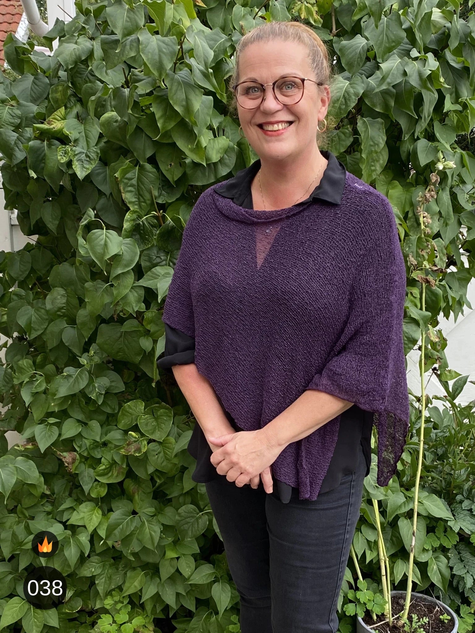 Kvinna bär Ella Cape i färg 038 Aubergine, en varm färg, finstickad cape som draperas mjukt över axlarna, fotad framför en grön häck. Ulla Jacobsson Kläder.