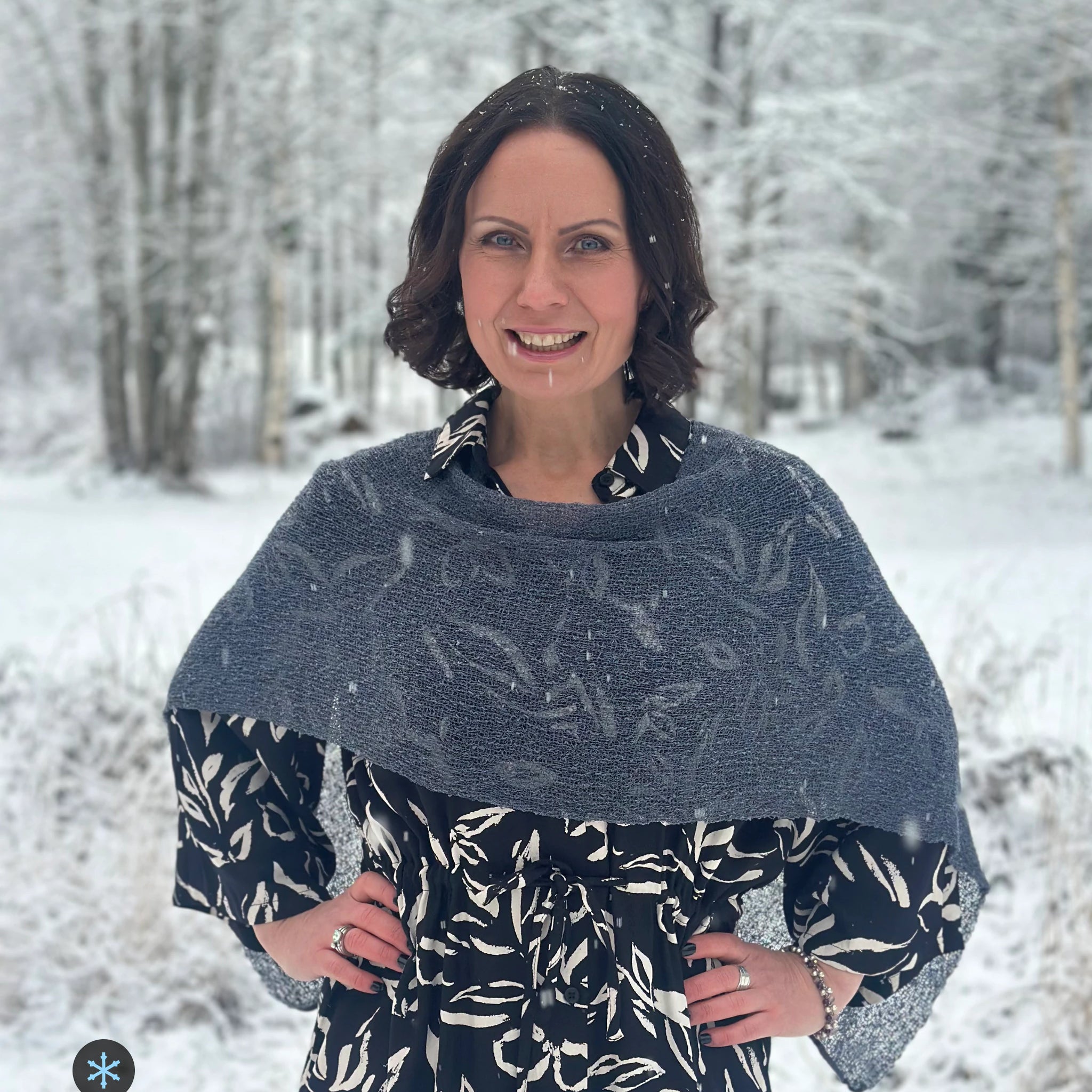 Kvinna bär Ella Cape i färg 039 Anilin, en kall färg i dov gråblå anilin, finstickad cape som draperas mjukt över axlarna, fotad i ett snöigt vinterlandskap. Ulla Jacobsson Kläder.