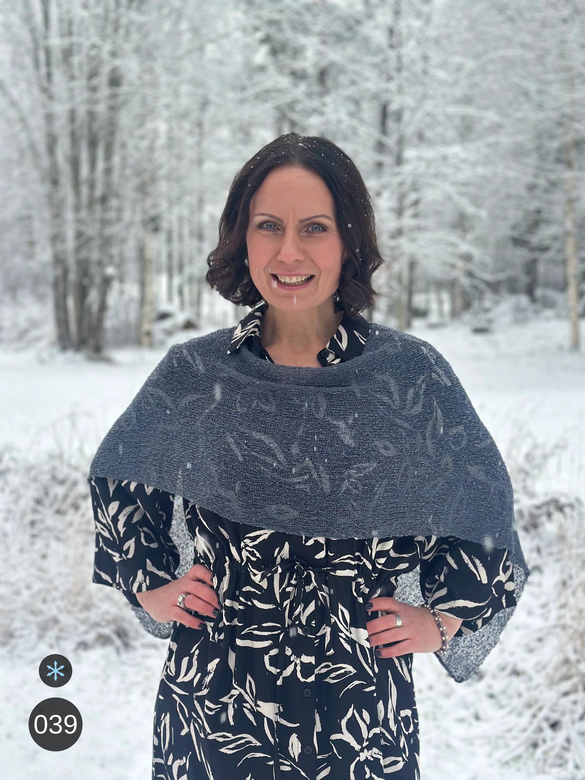 Kvinna bär Ella Cape i färg 039 Anilin, en kall färg i dov gråblå anilin, finstickad cape som draperas mjukt över axlarna, fotad i ett snöigt vinterlandskap. Ulla Jacobsson Kläder.