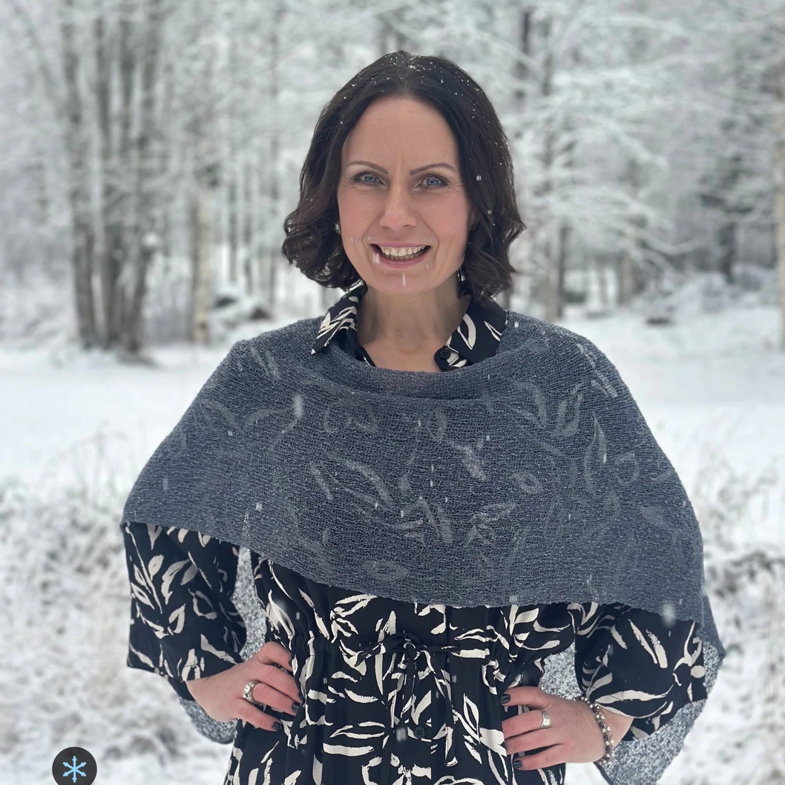 Kvinna bär Ella Cape i färg 039 Anilin, en kall färg, finstickad cape som draperas mjukt över axlarna, fotad i ett snöigt vinterlandskap. Ulla Jacobsson Kläder.