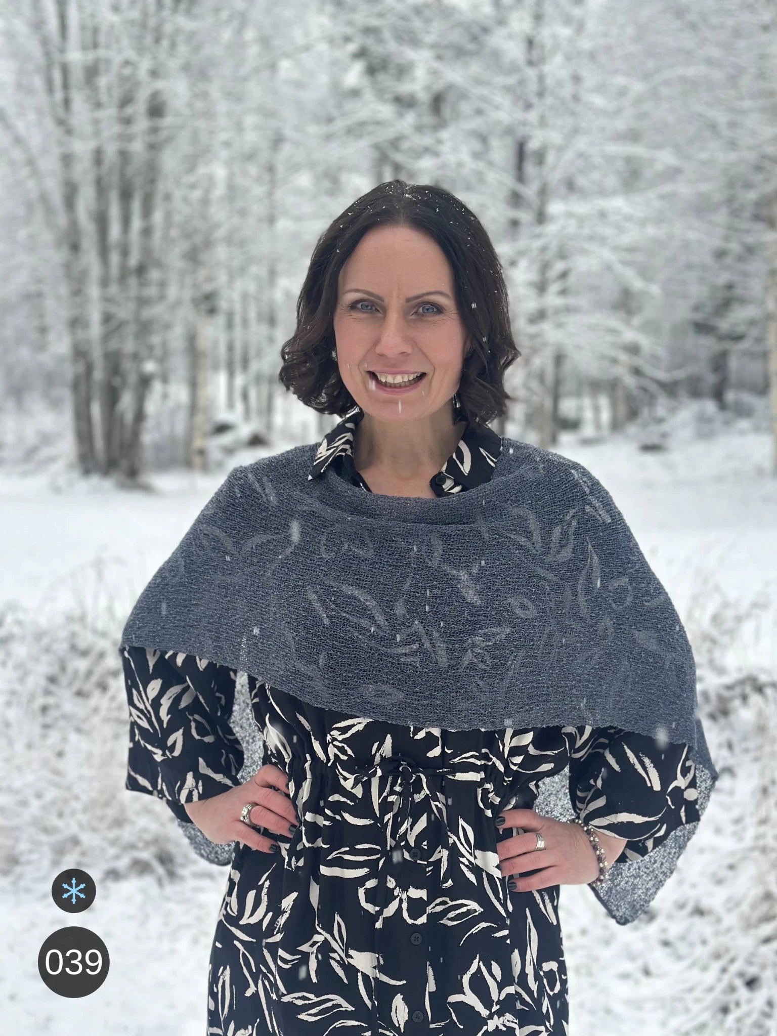 Kvinna bär Ella Cape i färg 039 Anilin, en kall färg, finstickad cape som draperas mjukt över axlarna, fotad i ett snöigt vinterlandskap. Ulla Jacobsson Kläder.
