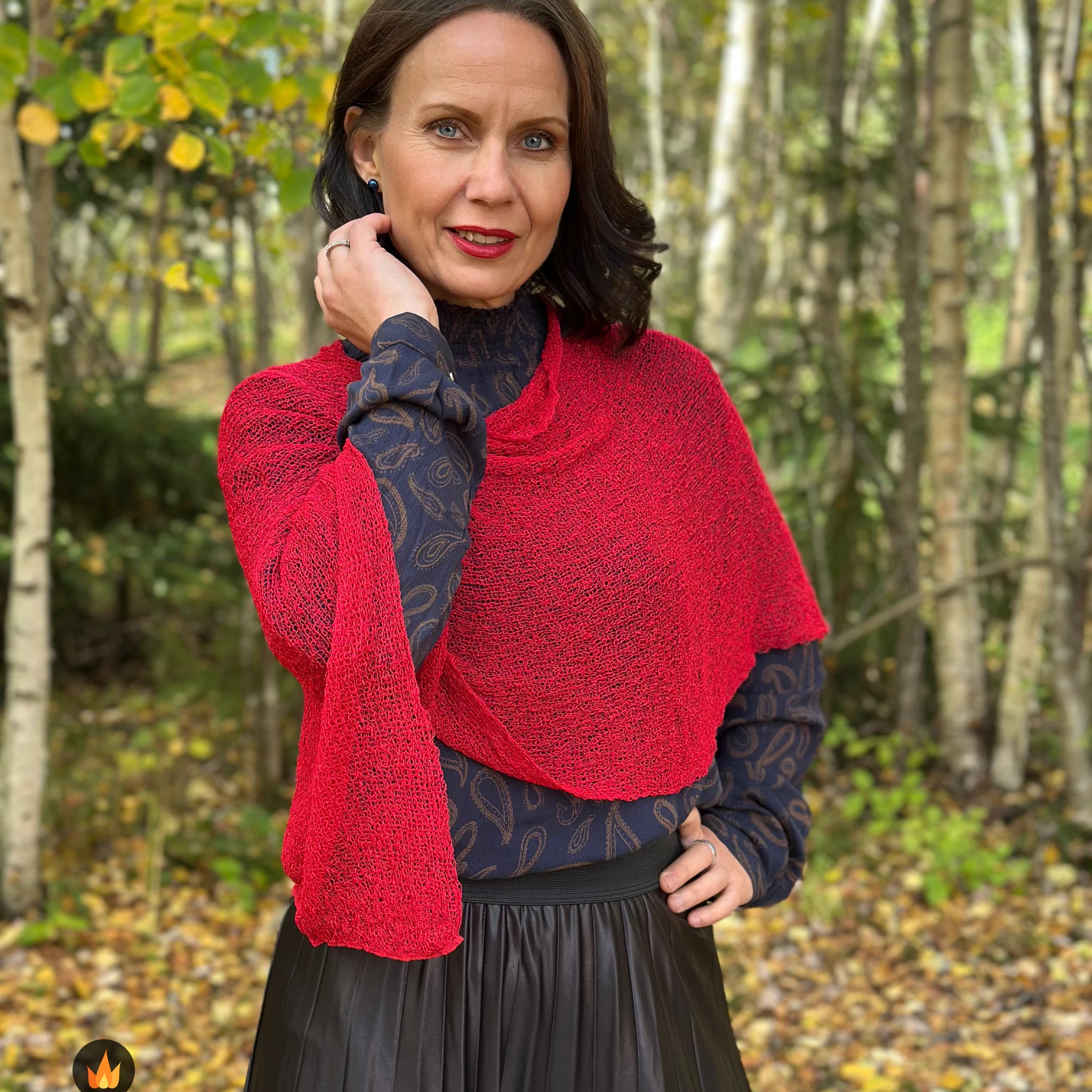 Kvinna bär Ella Cape i färg 042 Röd, en varm färg i klar röd med varm glöd, finstickad cape som draperas mjukt över axlarna, fotad bland höstgula björkar. Ulla Jacobsson Kläder.