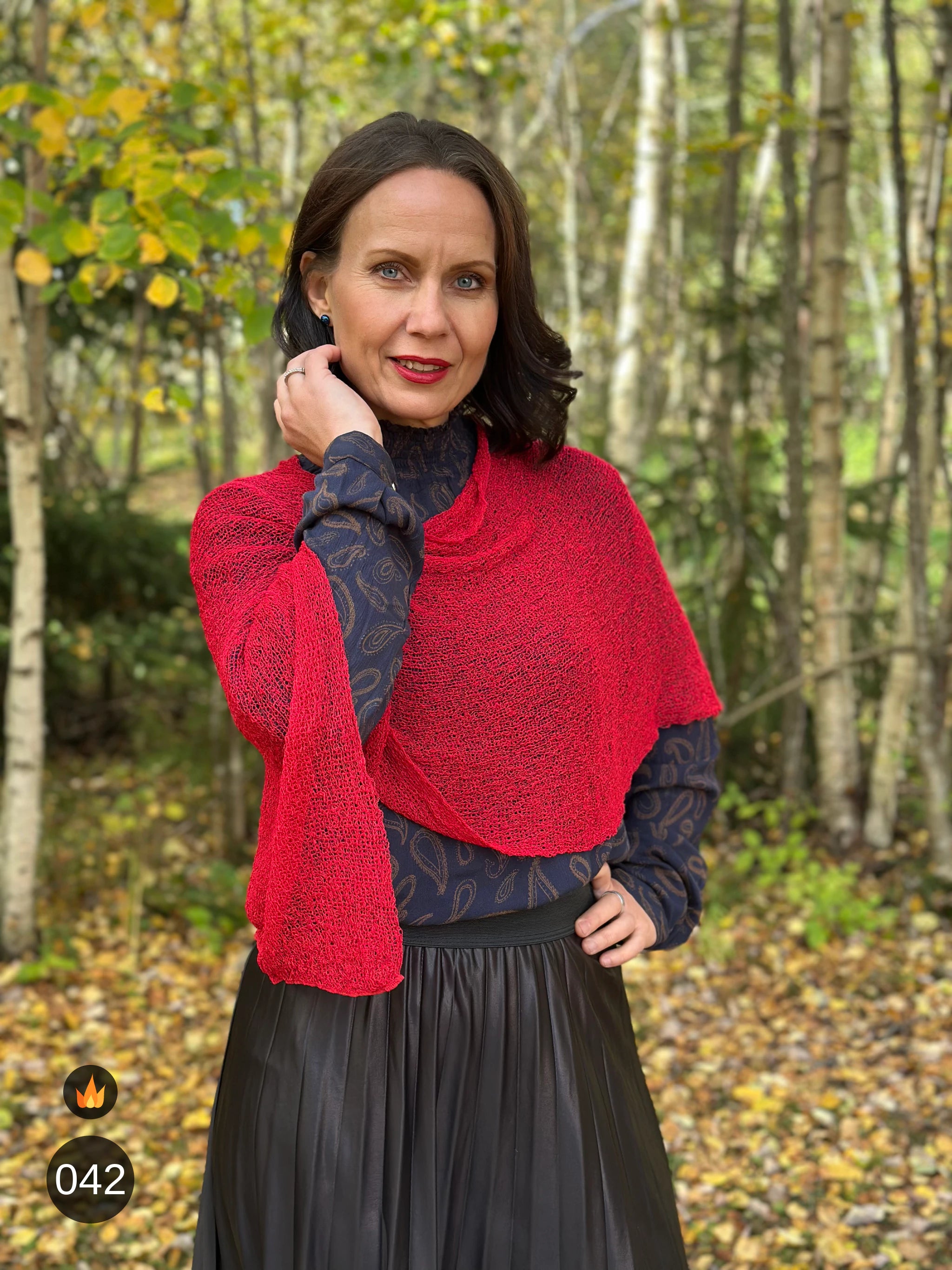 Kvinna bär Ella Cape i färg 042 Röd, en varm färg i klar röd med varm glöd, finstickad cape som draperas mjukt över axlarna, fotad bland höstgula björkar. Ulla Jacobsson Kläder.