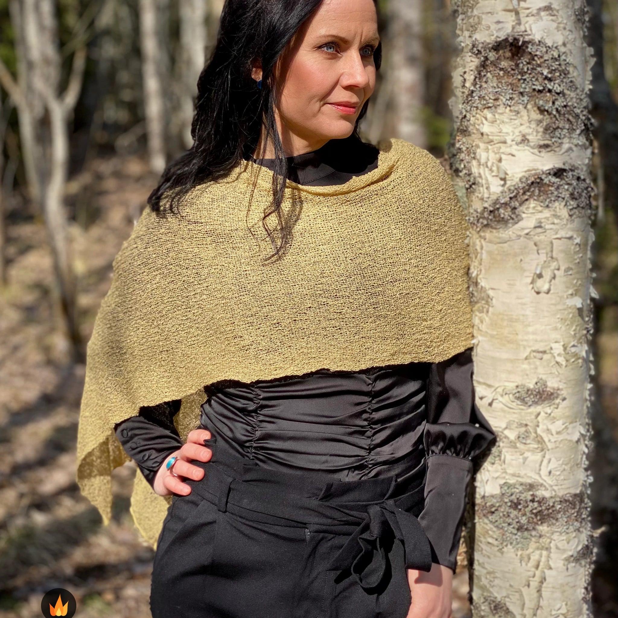 Kvinna bär Ella Cape i färg 046 Havre, en varm färg i havrebeige med gyllene ton, finstickad cape som draperas mjukt över axlarna, fotad vid en björkstam i skogsbrynet. Ulla Jacobsson Kläder.