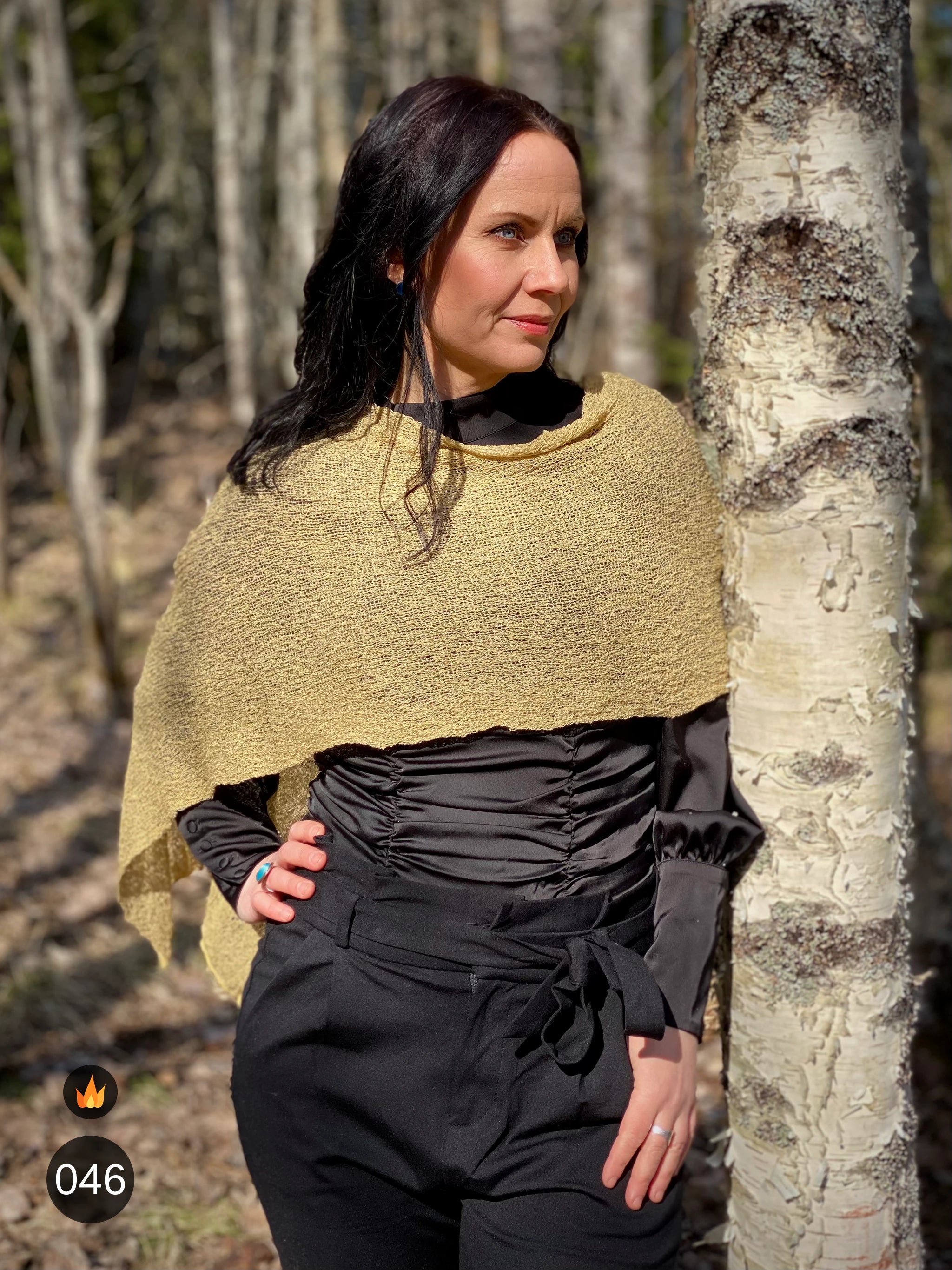 Kvinna bär Ella Cape i färg 046 Havre, en varm färg i havrebeige med gyllene ton, finstickad cape som draperas mjukt över axlarna, fotad vid en björkstam i skogsbrynet. Ulla Jacobsson Kläder.