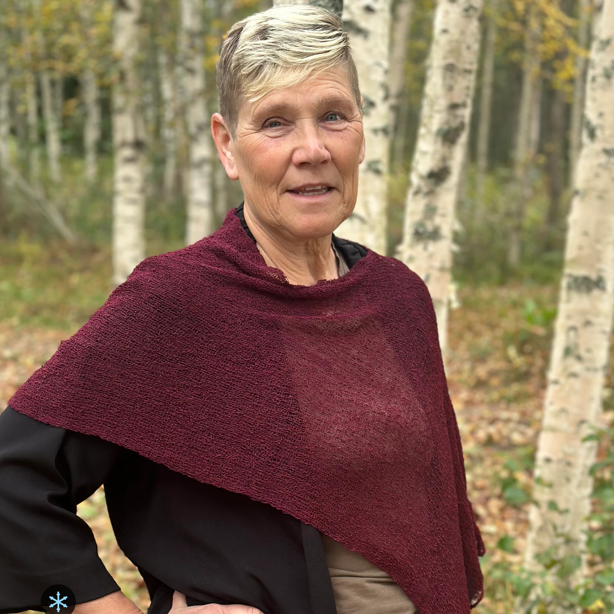 Kvinna bär Ella Cape i färg 047 Bourdeaux, en kall färg i djup bourdeauxröd med blå underton, finstickad cape som draperas mjukt över axlarna, fotad bland snöiga björkar. Ulla Jacobsson Kläder.