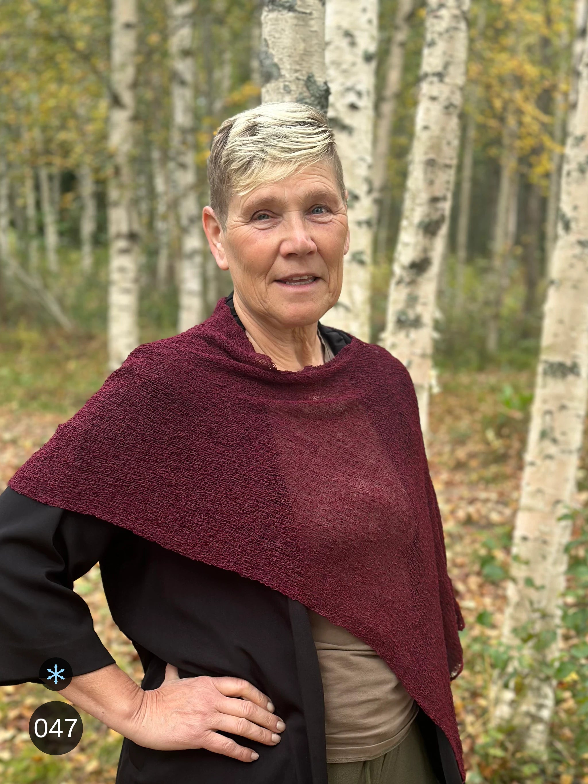 Kvinna bär Ella Cape i färg 047 Bourdeaux, en kall färg i djup bourdeauxröd med blå underton, finstickad cape som draperas mjukt över axlarna, fotad bland snöiga björkar. Ulla Jacobsson Kläder.