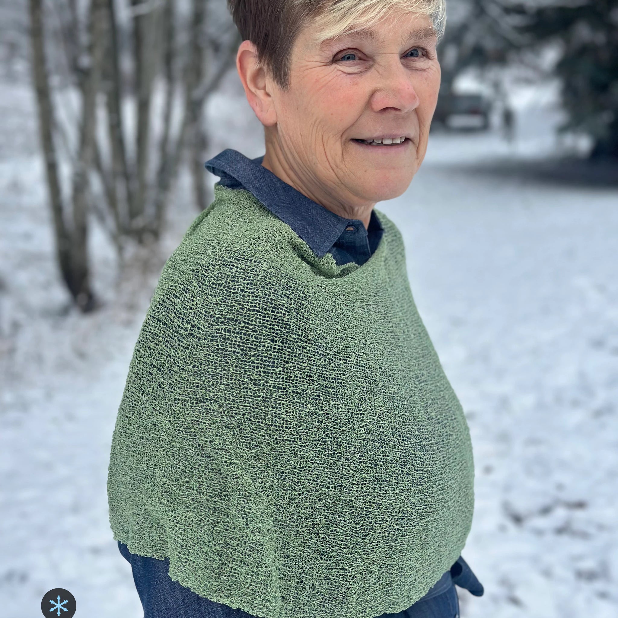 Kvinna bär Ella Cape i färg 050 Skogsgrön, en kall färg i dämpad skogsgrön, finstickad cape som draperas mjukt över axlarna, fotad i en frostig allé. Ulla Jacobsson Kläder.