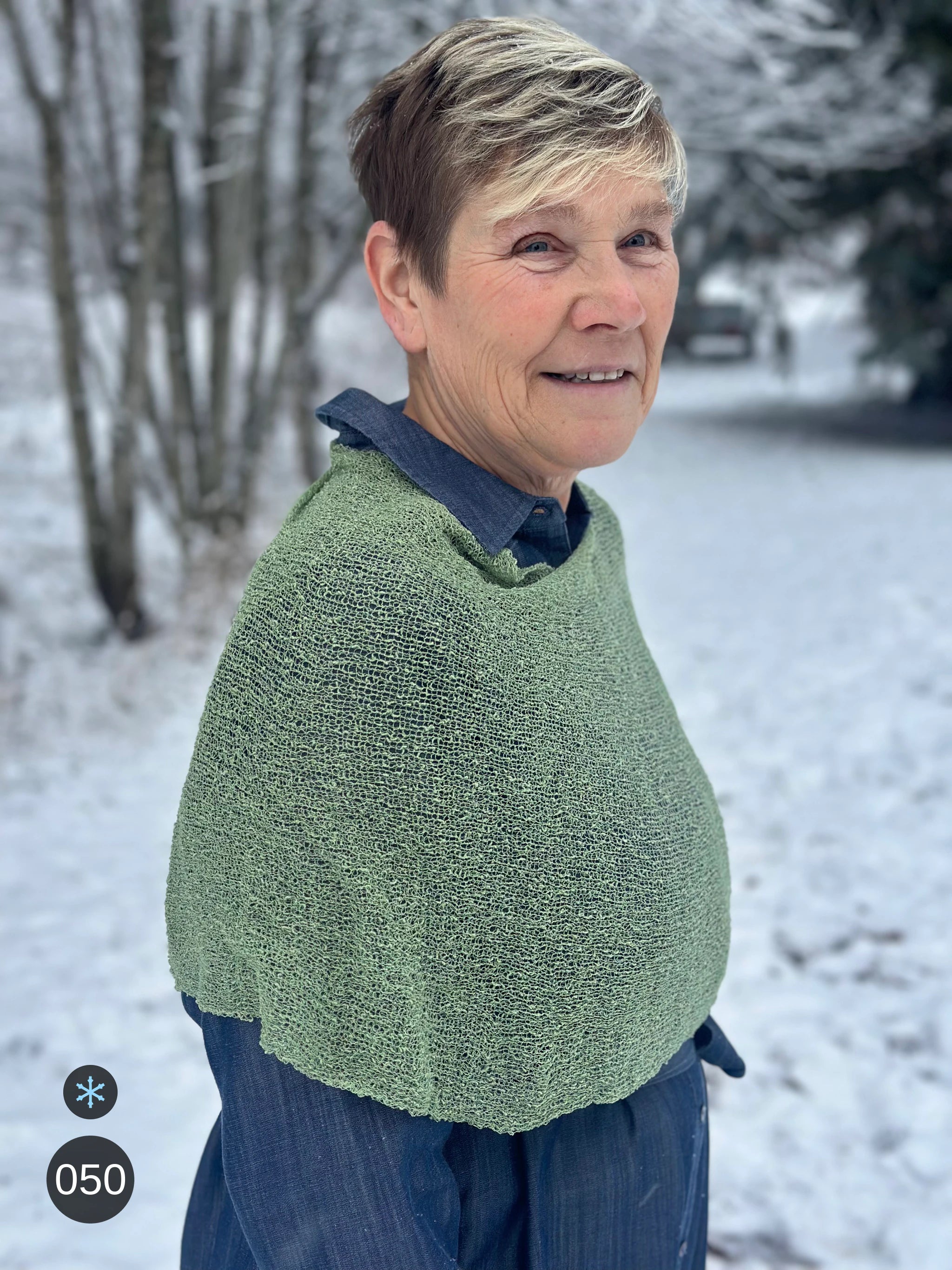 Kvinna bär Ella Cape i färg 050 Skogsgrön, en kall färg i dämpad skogsgrön, finstickad cape som draperas mjukt över axlarna, fotad i en frostig allé. Ulla Jacobsson Kläder.
