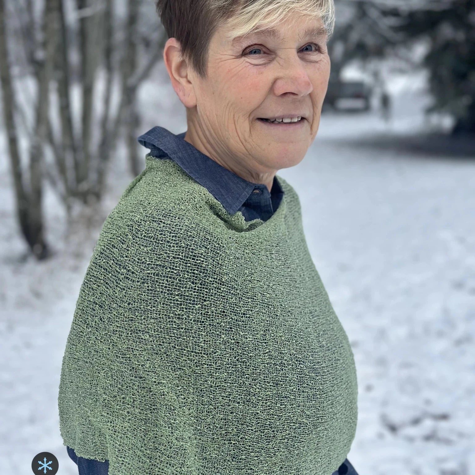 Kvinna bär Ella Cape i färg 050 Skogsgrön, en kall färg, finstickad cape som draperas mjukt över axlarna, fotad i en frostig allé. Ulla Jacobsson Kläder.