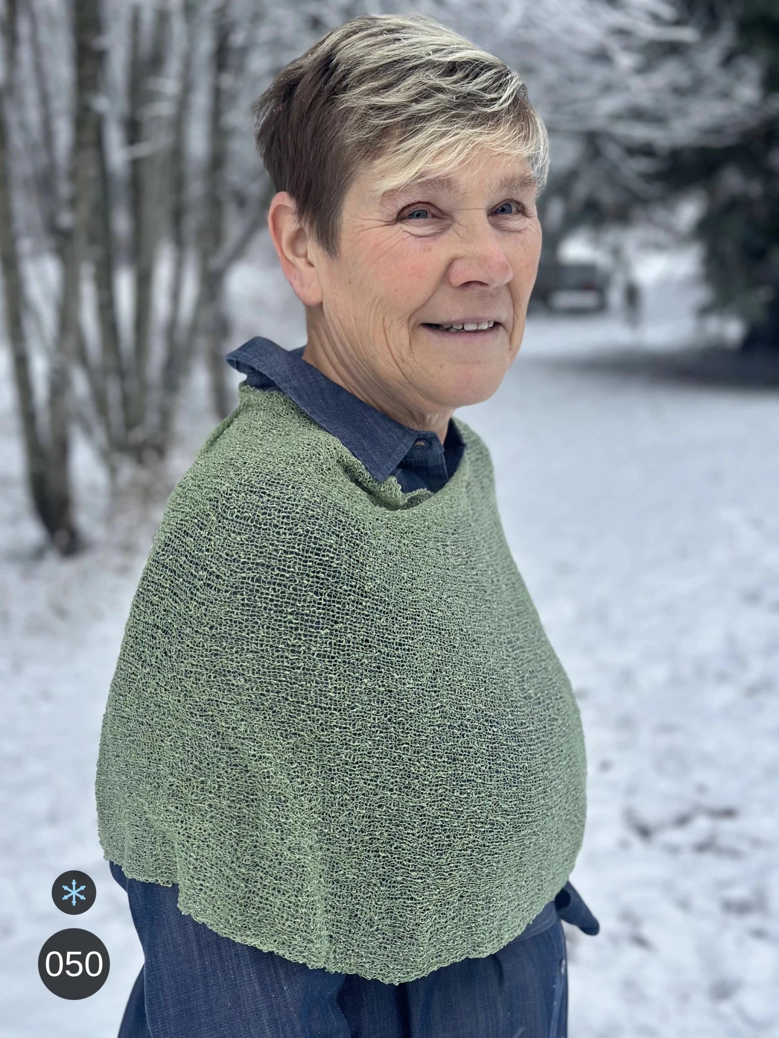 Kvinna bär Ella Cape i färg 050 Skogsgrön, en kall färg, finstickad cape som draperas mjukt över axlarna, fotad i en frostig allé. Ulla Jacobsson Kläder.