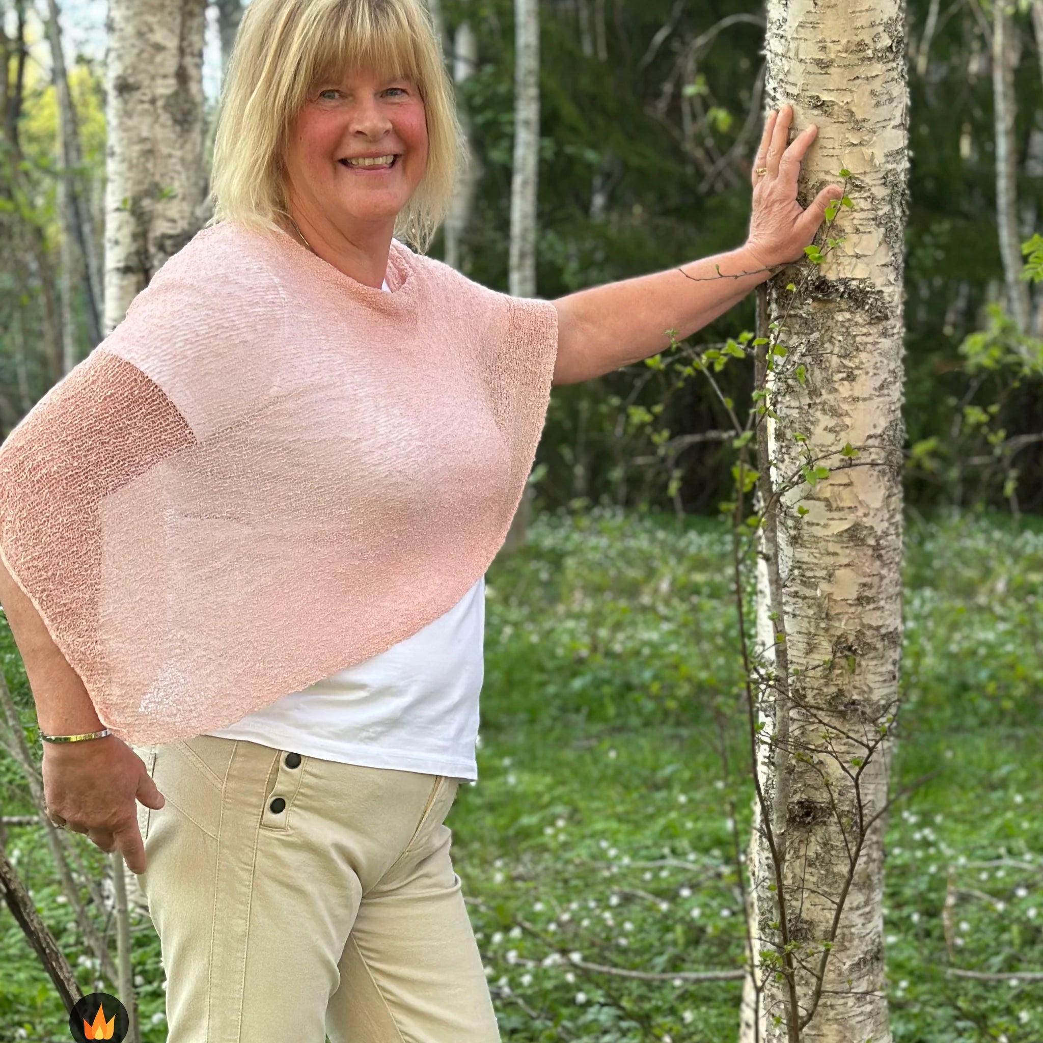 Kvinna bär Ella Cape i färg 051 Puderrosa, en varm färg i ljus puderrosa, finstickad cape som draperas mjukt över axlarna, fotad i en björkdunge en försommardag. Ulla Jacobsson Kläder.