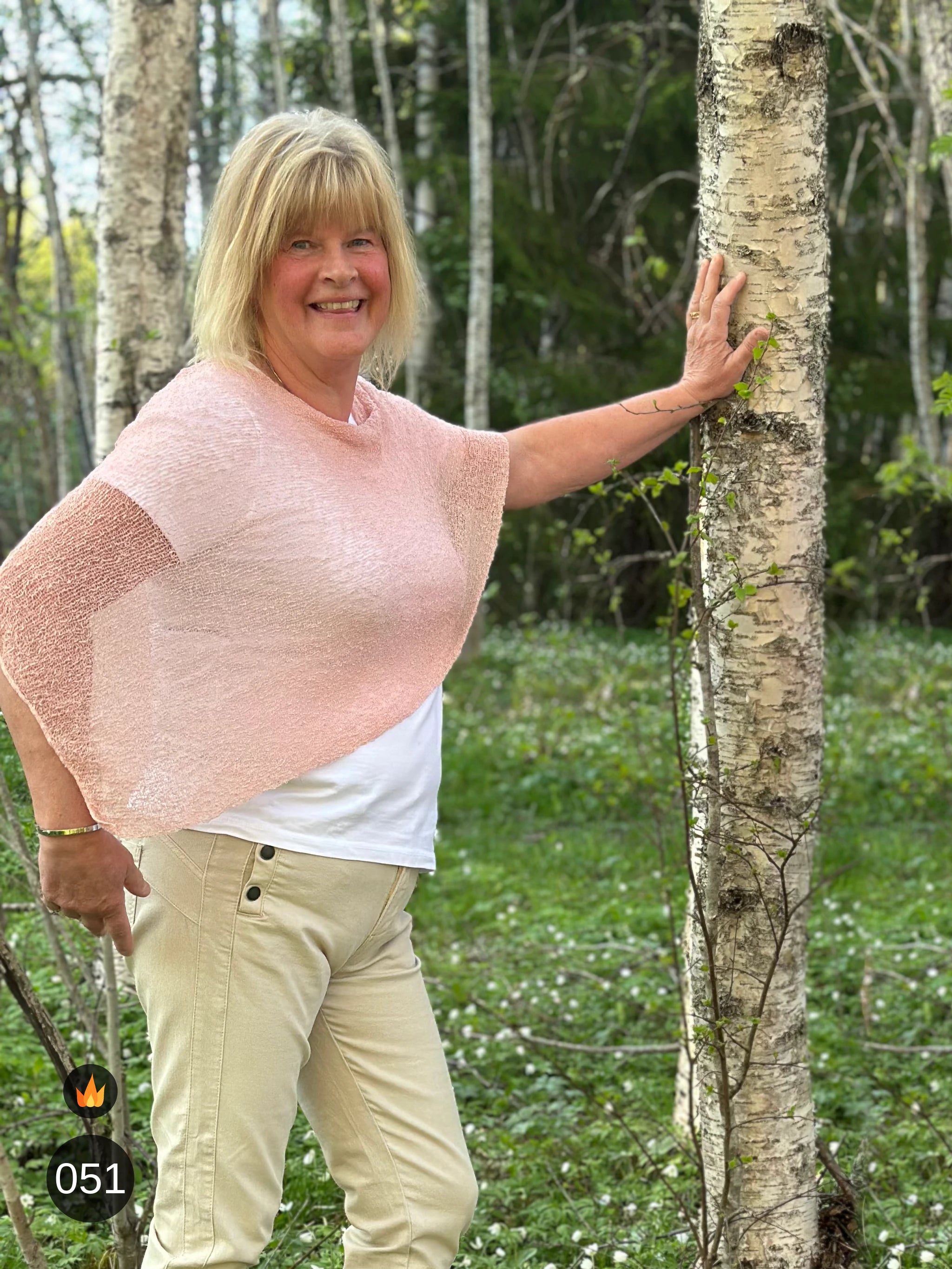 Kvinna bär Ella Cape i färg 051 Puderrosa, en varm färg i ljus puderrosa, finstickad cape som draperas mjukt över axlarna, fotad i en björkdunge en försommardag. Ulla Jacobsson Kläder.