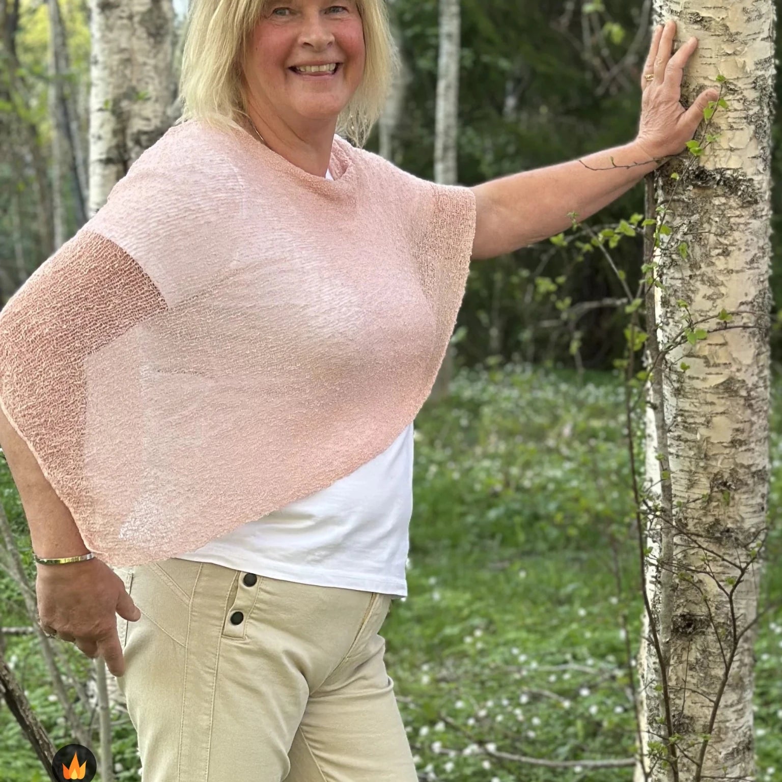 Kvinna bär Ella Cape i färg 051 Puderrosa, en varm färg, finstickad cape som draperas mjukt över axlarna, fotad vid en björkstam en försommardag. Ulla Jacobsson Kläder.