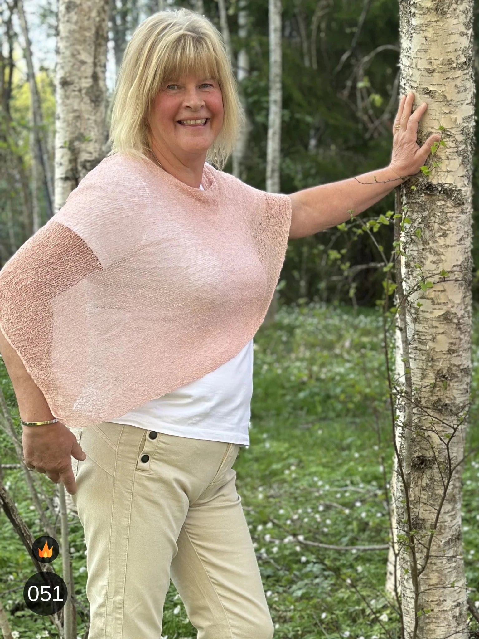 Kvinna bär Ella Cape i färg 051 Puderrosa, en varm färg, finstickad cape som draperas mjukt över axlarna, fotad vid en björkstam en försommardag. Ulla Jacobsson Kläder.