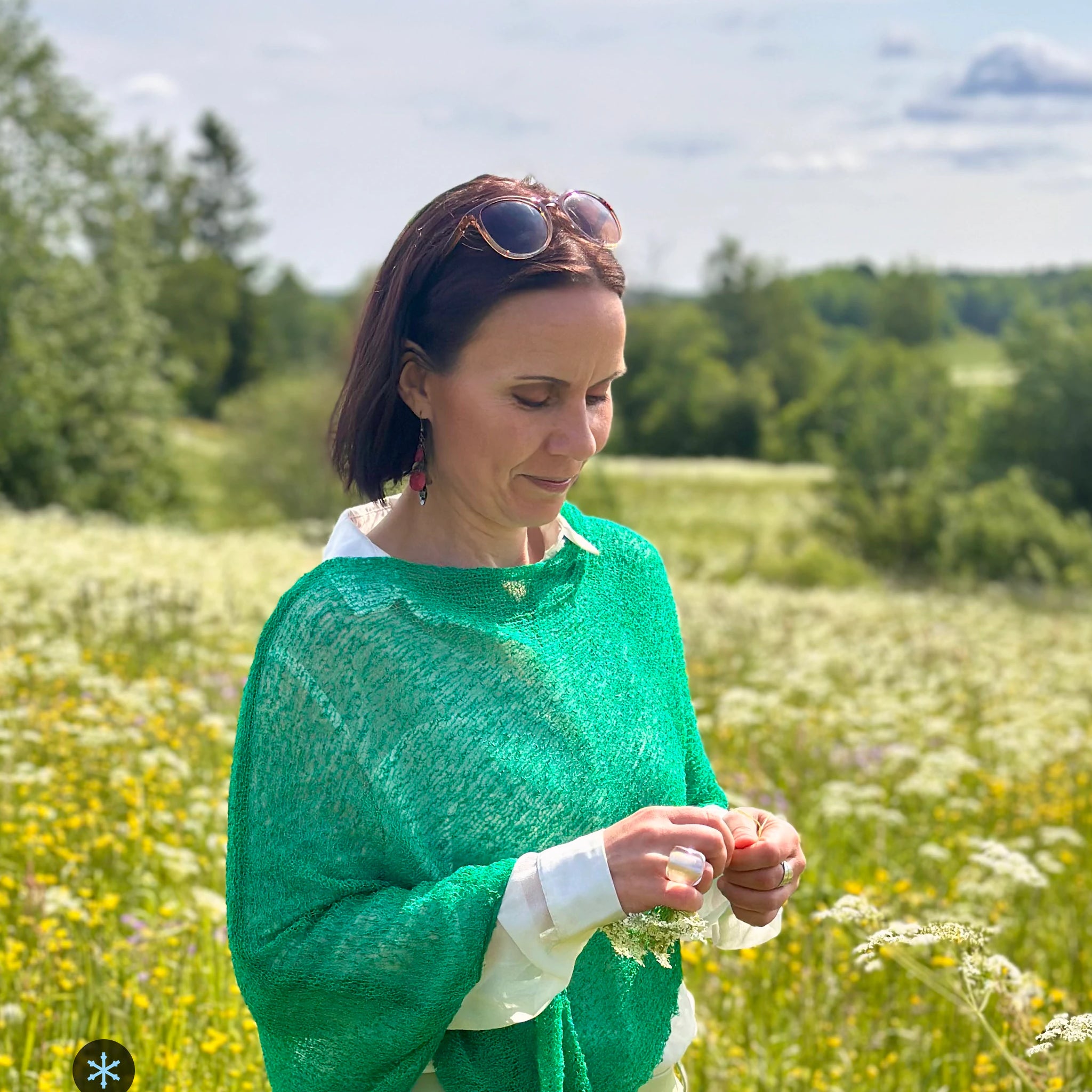 Kvinna bär Ella Cape i färg 052 Bladgrön, en kall färg i klar bladgrön, finstickad cape som draperas mjukt över axlarna, fotad på en blommande sommaräng. Ulla Jacobsson Kläder.
