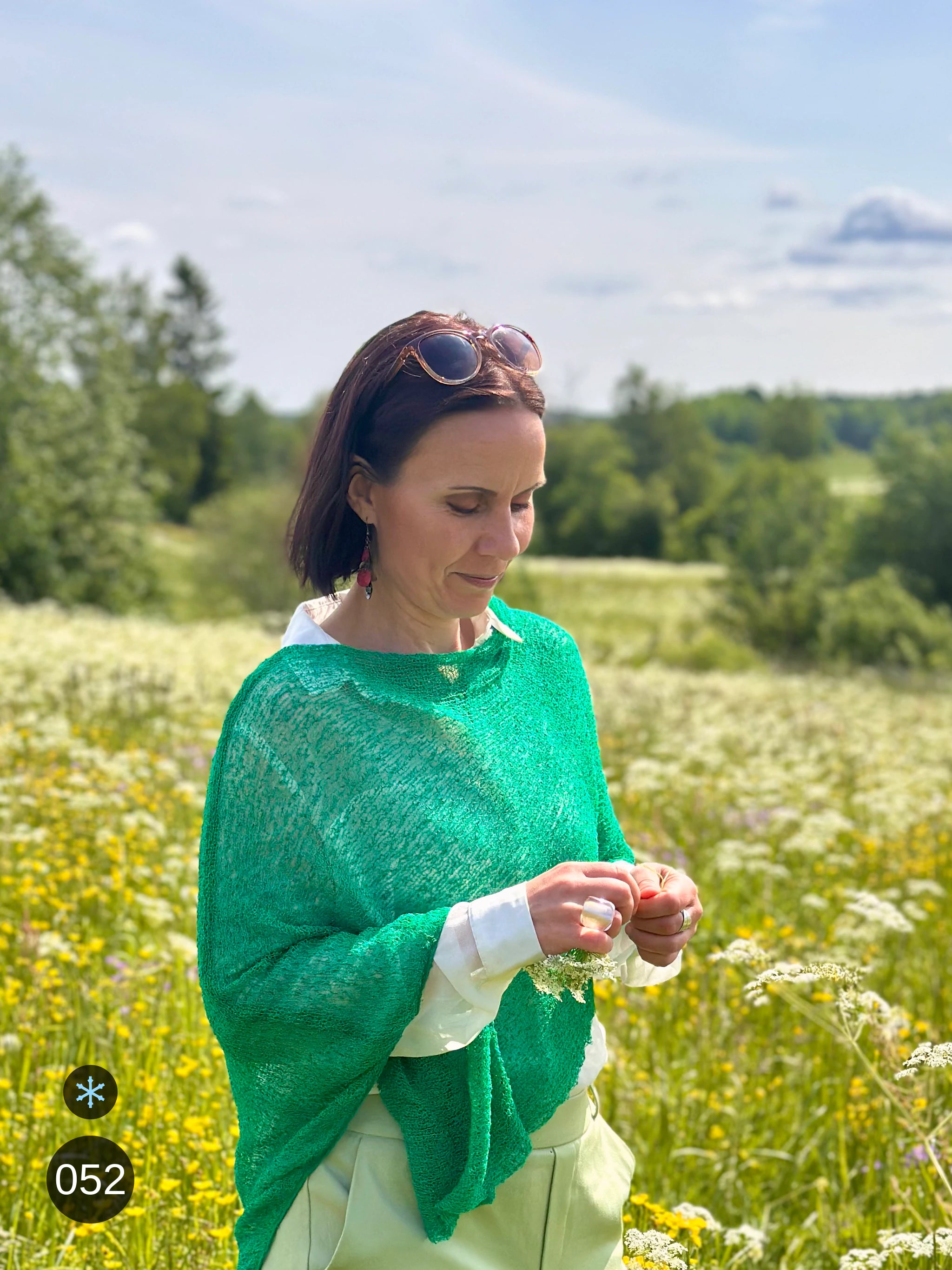 Kvinna bär Ella Cape i färg 052 Bladgrön, en kall färg i klar bladgrön, finstickad cape som draperas mjukt över axlarna, fotad på en blommande sommaräng. Ulla Jacobsson Kläder.