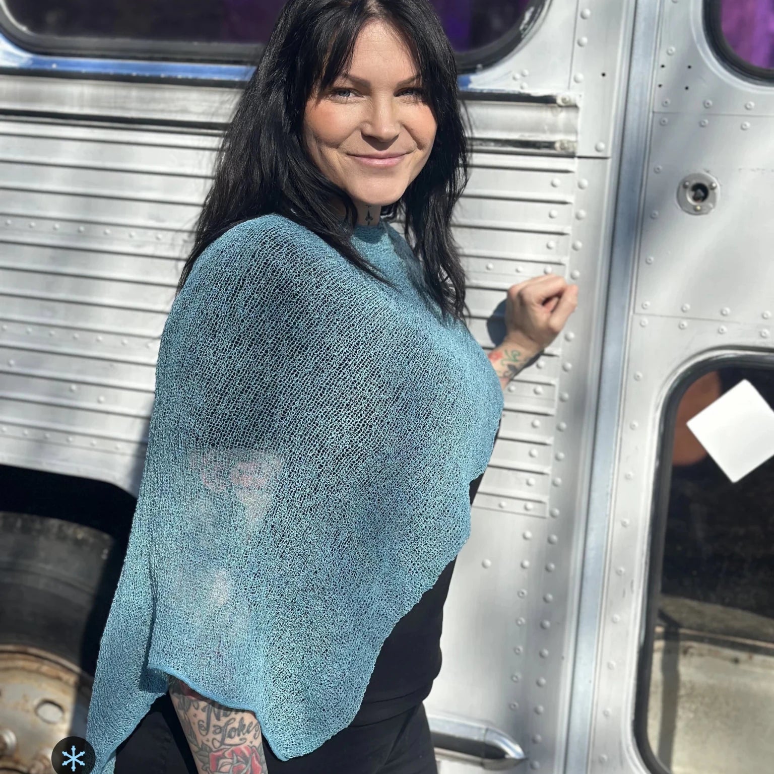 Kvinna bär Ella Cape i färg 053 Aqua, en kall färg, finstickad cape som draperas mjukt över axlarna, fotad utomhus framför en silverfärgad veteranbuss. Ulla Jacobsson Kläder.