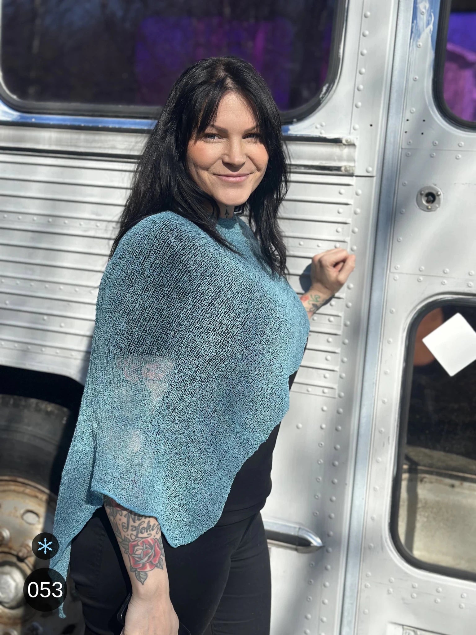 Kvinna bär Ella Cape i färg 053 Aqua, en kall färg, finstickad cape som draperas mjukt över axlarna, fotad utomhus framför en silverfärgad veteranbuss. Ulla Jacobsson Kläder.