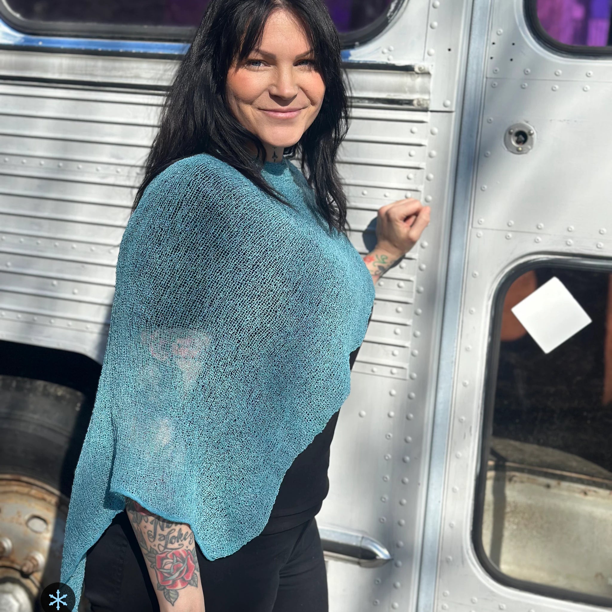 Kvinna bär Ella Cape i färg 053 Aqua, en kall färg i klar aqua med grön skiftning, finstickad cape som draperas mjukt över axlarna, fotad framför en silverfärgad veteranbuss i vårsol. Ulla Jacobsson Kläder.