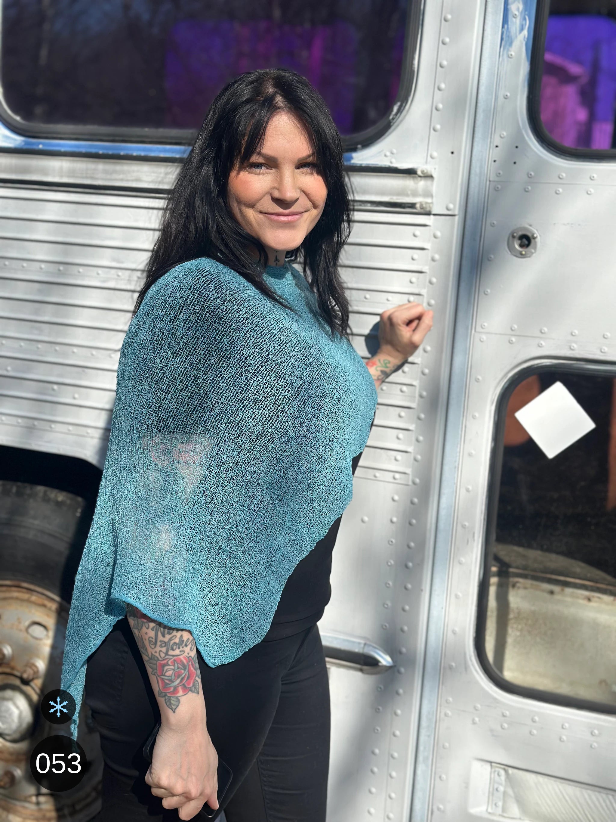 Kvinna bär Ella Cape i färg 053 Aqua, en kall färg i klar aqua med grön skiftning, finstickad cape som draperas mjukt över axlarna, fotad framför en silverfärgad veteranbuss i vårsol. Ulla Jacobsson Kläder.