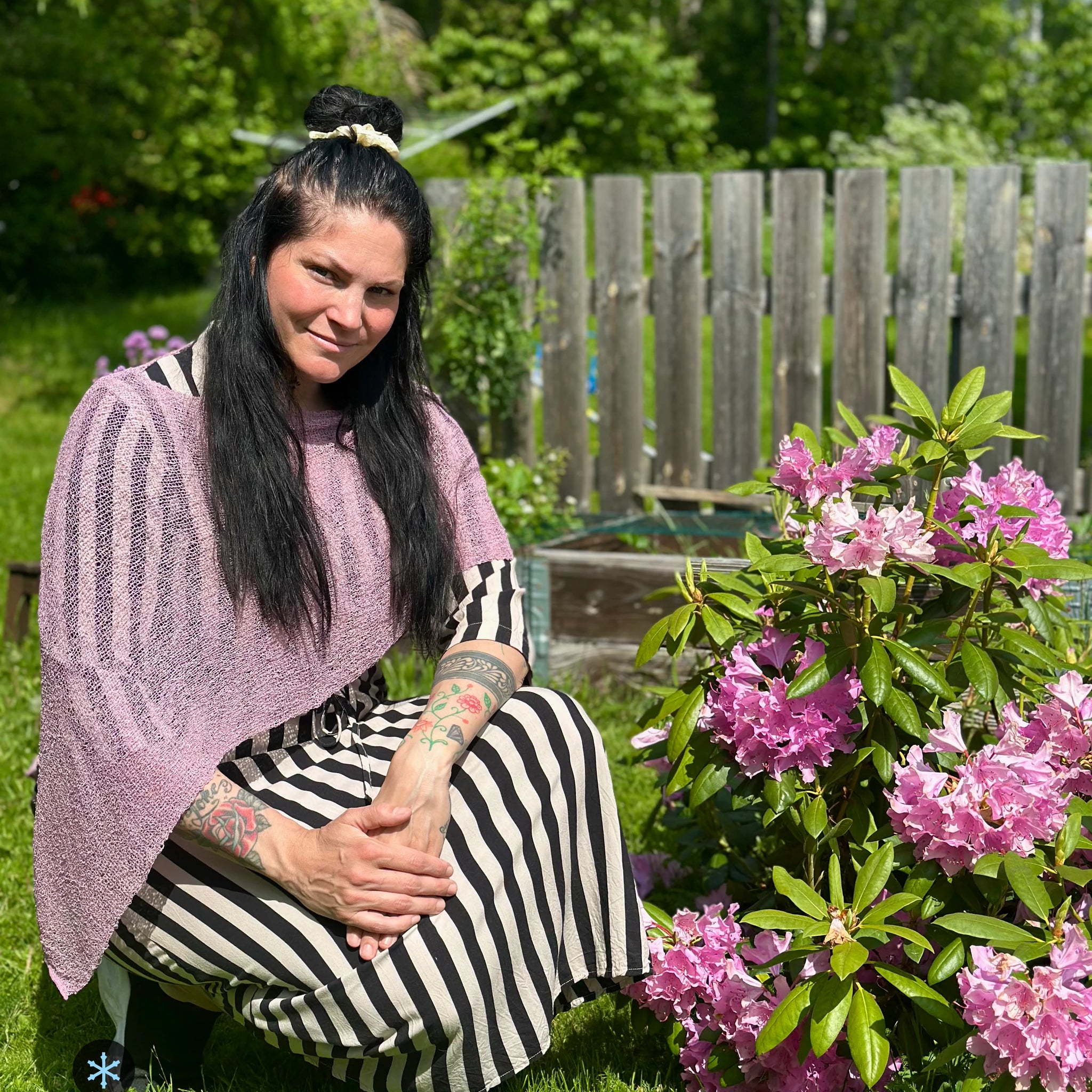Kvinna bär Ella Cape i färg 057 Lingon och mjölk, en kall färg i ljus lingonrosa med mjölkvit skiftning, finstickad cape som draperas mjukt över axlarna, fotad vid blommande rododendron. Ulla Jacobsson Kläder.