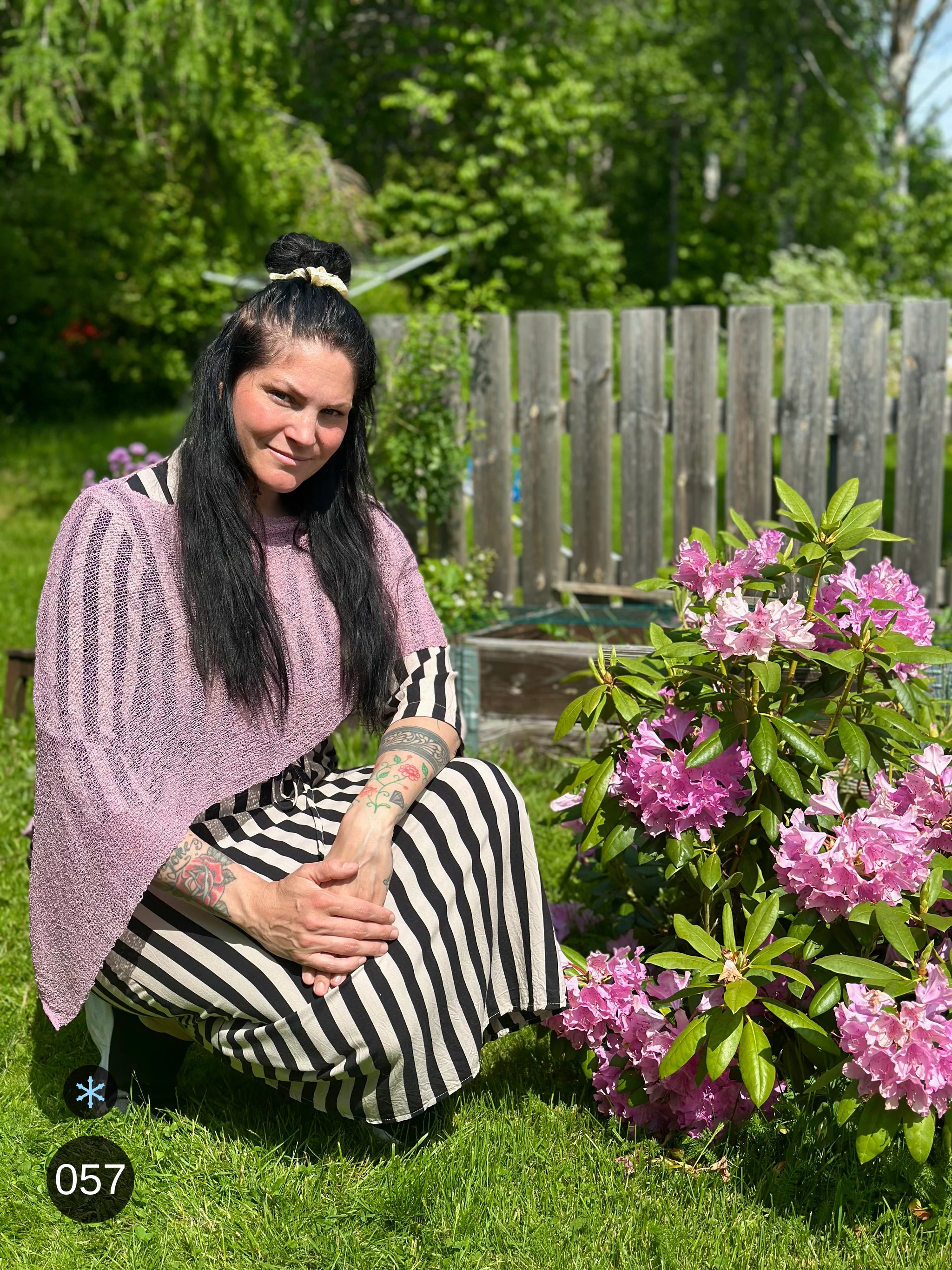 Kvinna bär Ella Cape i färg 057 Lingon och mjölk, en kall färg i ljus lingonrosa med mjölkvit skiftning, finstickad cape som draperas mjukt över axlarna, fotad vid blommande rododendron. Ulla Jacobsson Kläder.