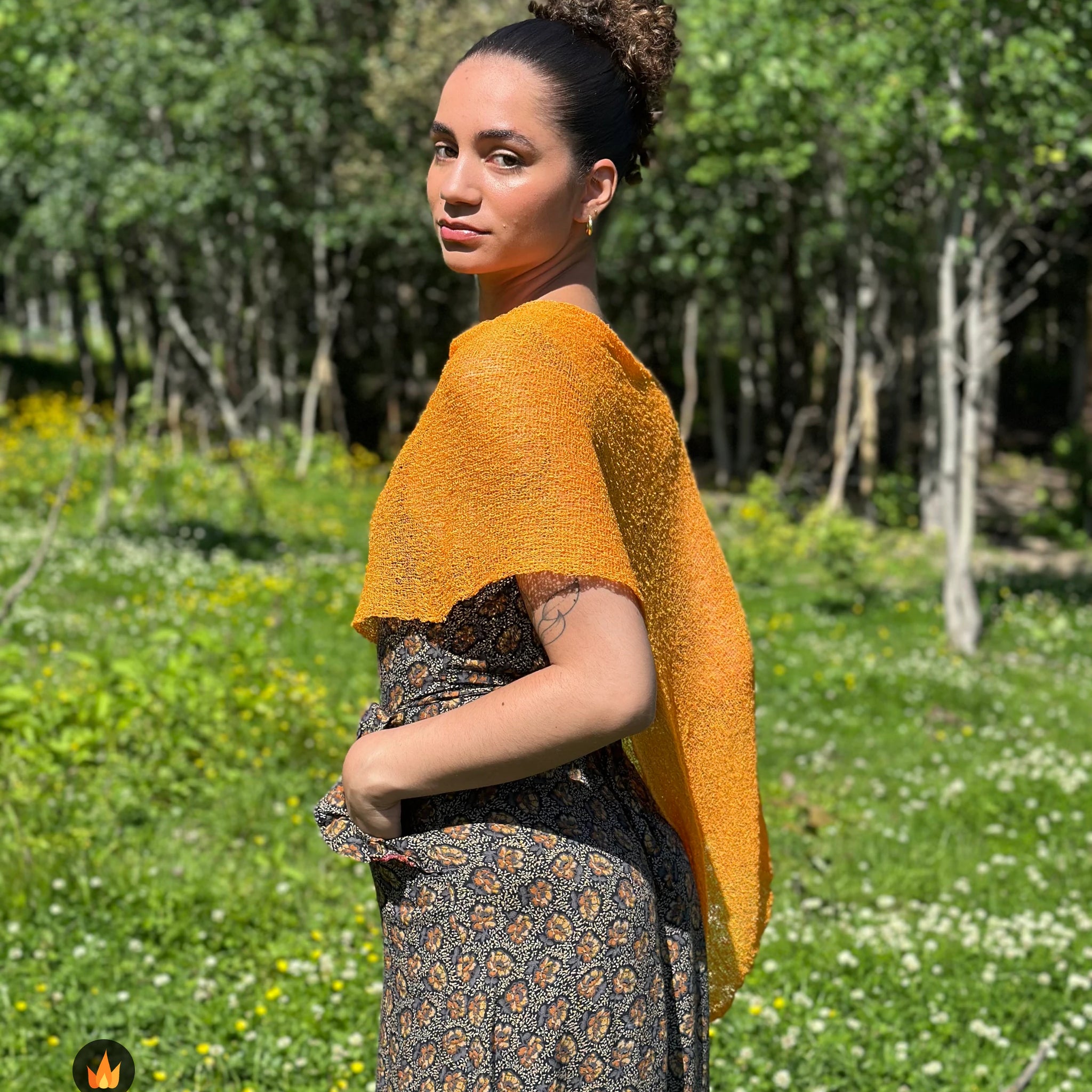 Kvinna bär Ella Cape i färg 059 Ljusorange, en varm färg i ljus orange med gyllene ton, finstickad cape som draperas mjukt över axlarna, fotad i försommargrönska. Ulla Jacobsson Kläder.