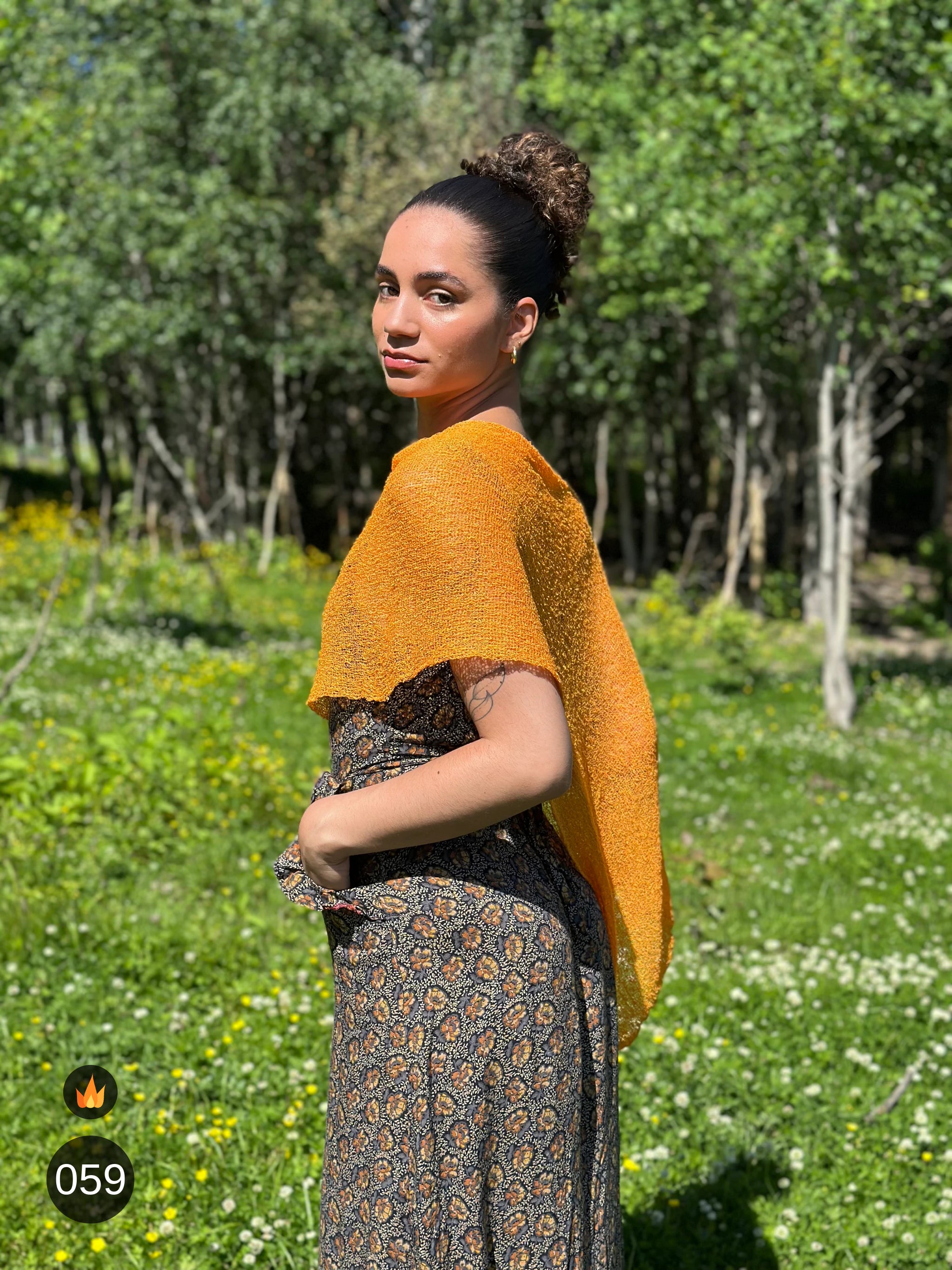 Kvinna bär Ella Cape i färg 059 Ljusorange, en varm färg i ljus orange med gyllene ton, finstickad cape som draperas mjukt över axlarna, fotad i försommargrönska. Ulla Jacobsson Kläder.