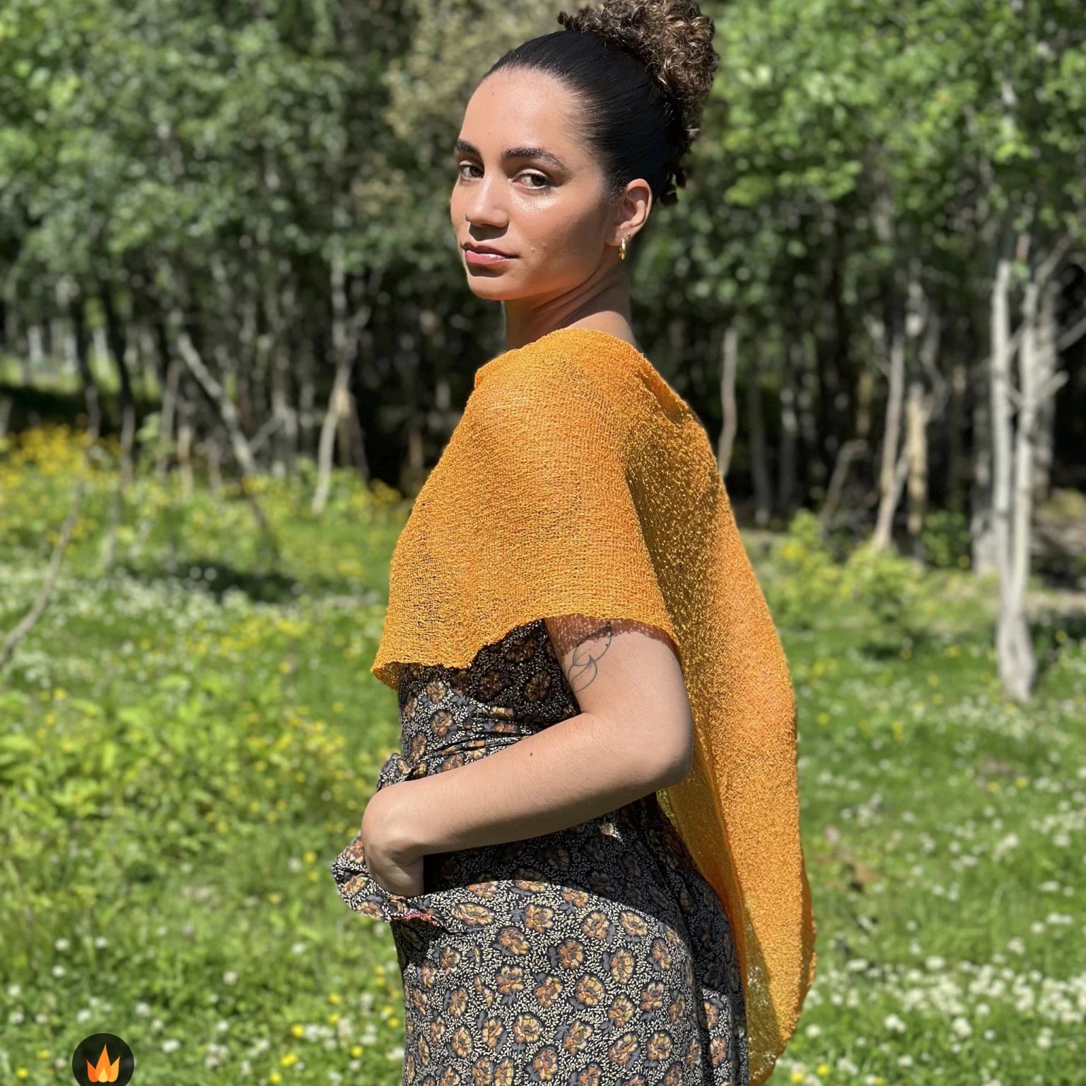 Kvinna bär Ella Cape i färg 059 Ljusorange, en varm färg, finstickad cape som draperas mjukt över axlarna, fotad i försommargrönska. Ulla Jacobsson Kläder.