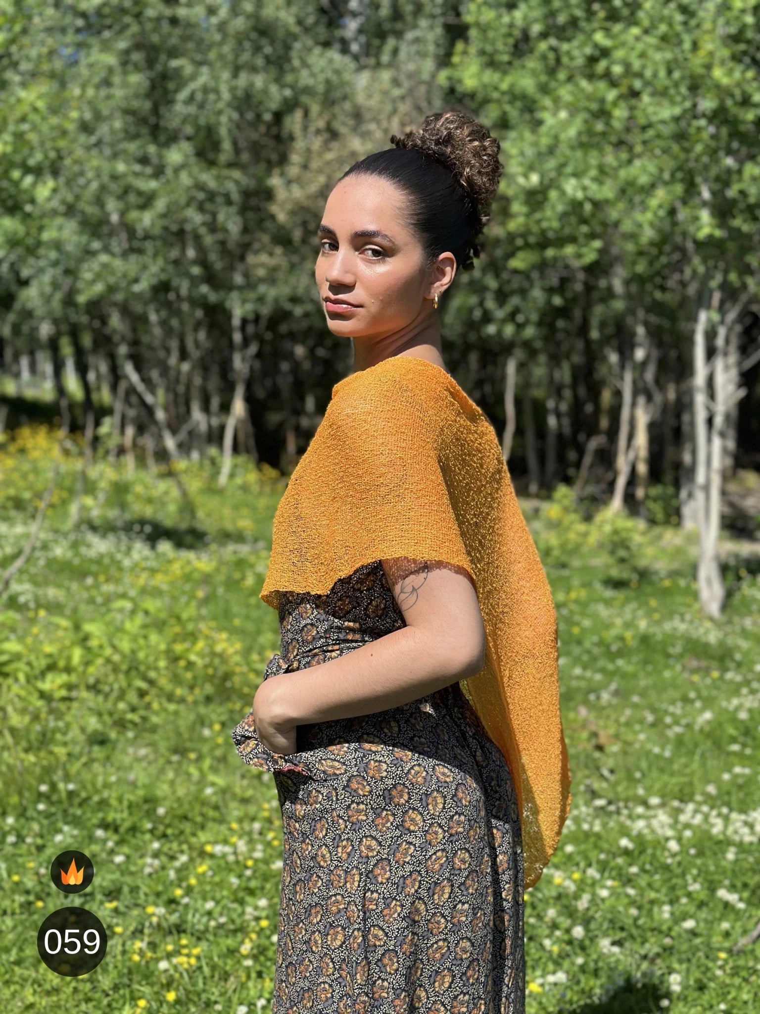 Kvinna bär Ella Cape i färg 059 Ljusorange, en varm färg, finstickad cape som draperas mjukt över axlarna, fotad i försommargrönska. Ulla Jacobsson Kläder.
