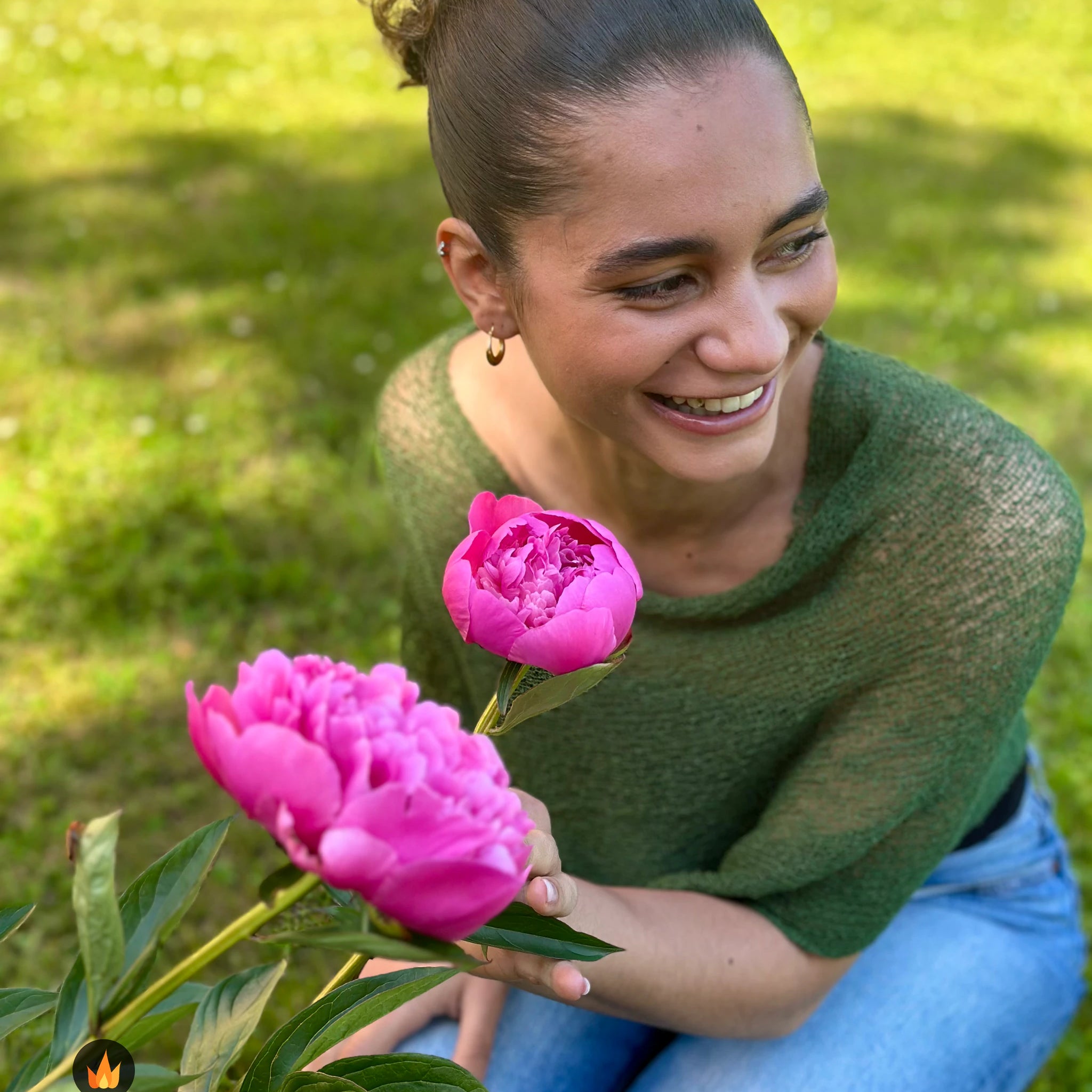 Kvinna bär Ella Cape i färg 061 Mörkgrön, en varm färg i djup mörkgrön, finstickad cape som draperas mjukt över axlarna, fotad i en trädgård med en rosa pion i handen. Ulla Jacobsson Kläder.