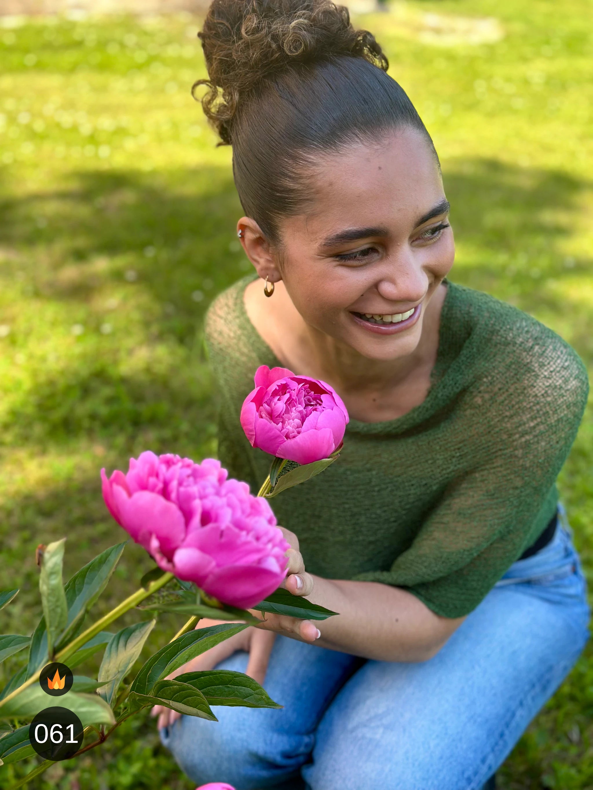 Kvinna bär Ella Cape i färg 061 Mörkgrön, en varm färg i djup mörkgrön, finstickad cape som draperas mjukt över axlarna, fotad i en trädgård med en rosa pion i handen. Ulla Jacobsson Kläder.