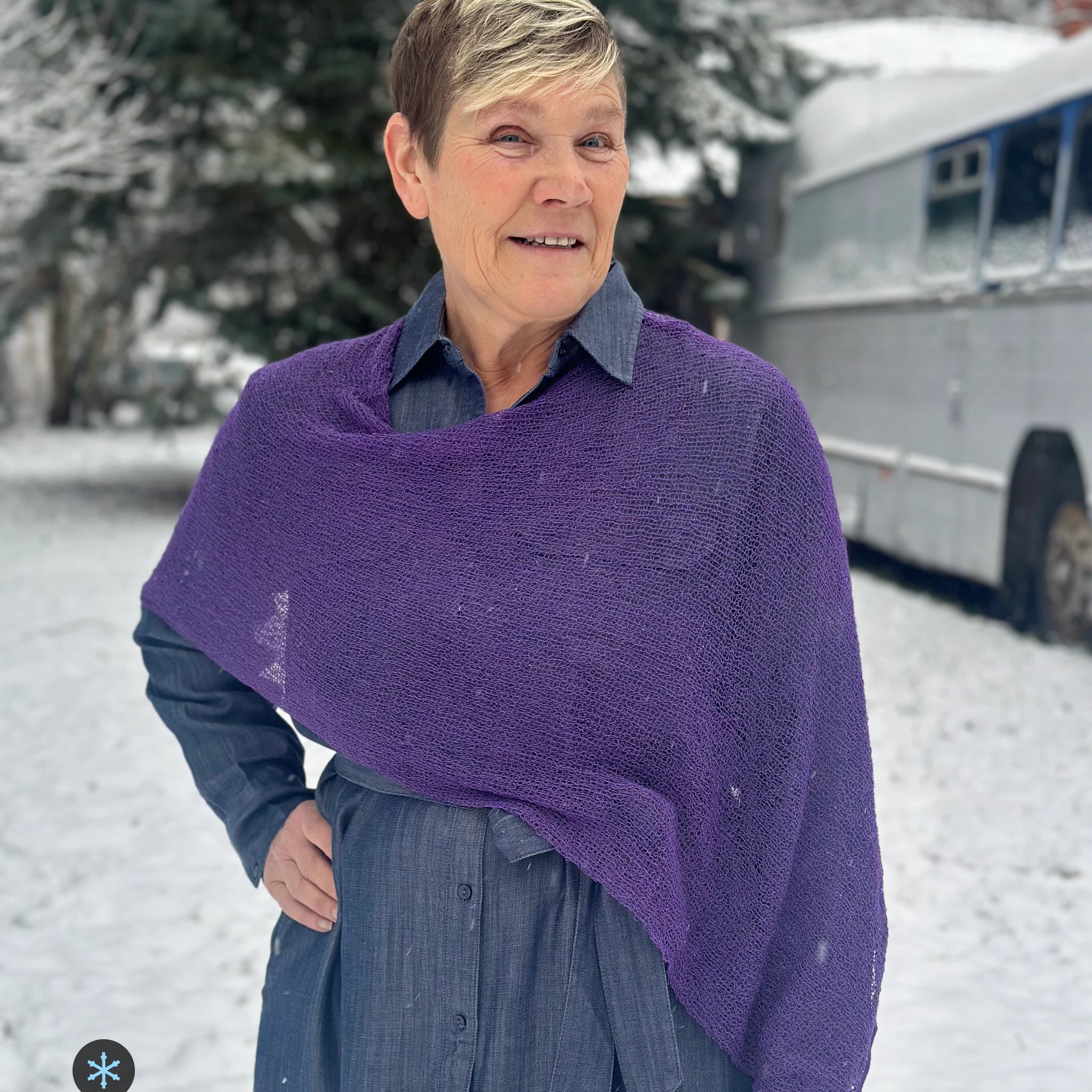 Kvinna bär Ella Cape i färg 065 Plommonlila, en kall färg i dov plommonlila, finstickad cape som draperas mjukt över axlarna, fotad i snö framför en silverfärgad veteranbuss. Ulla Jacobsson Kläder.