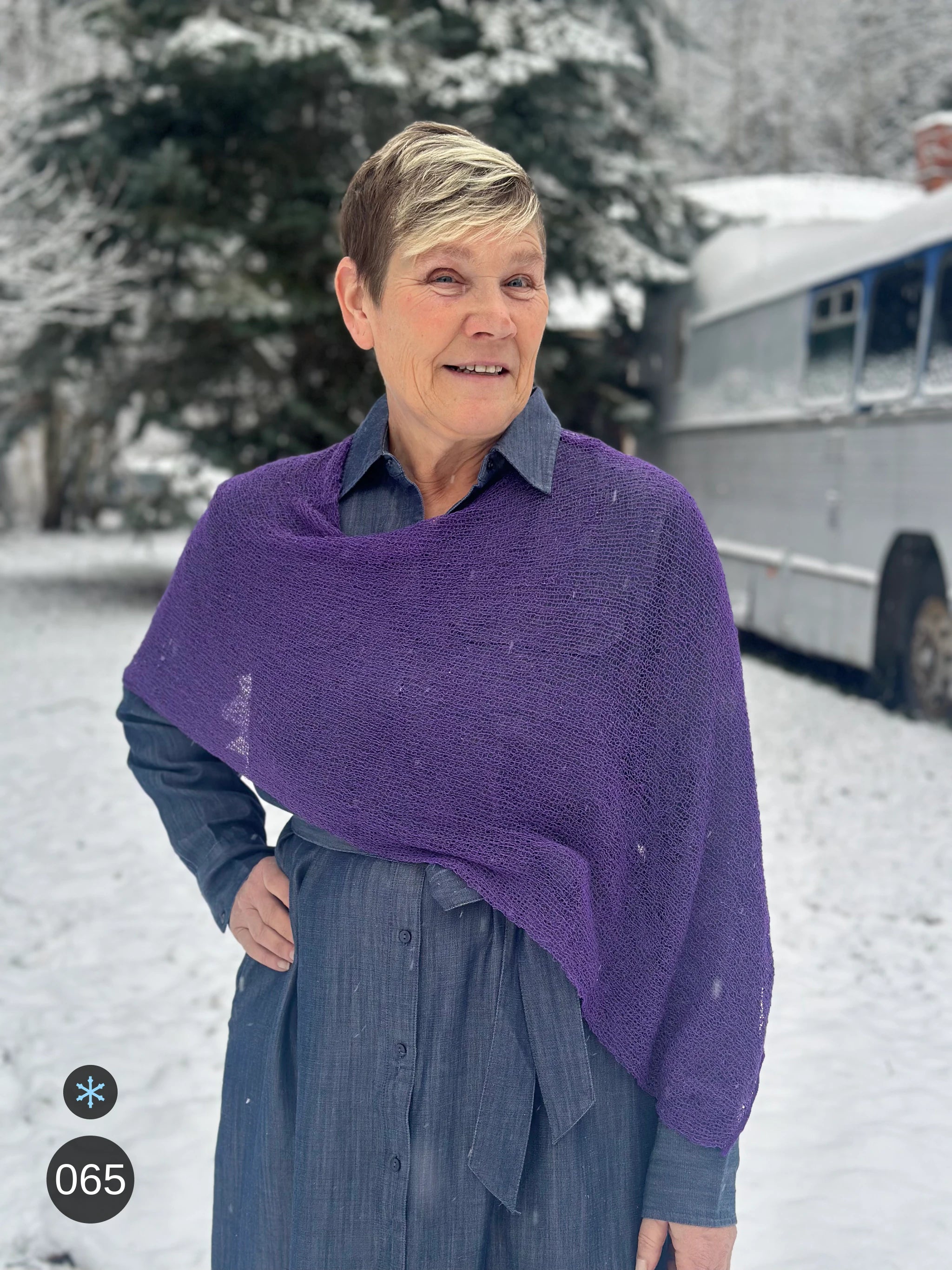 Kvinna bär Ella Cape i färg 065 Plommonlila, en kall färg i dov plommonlila, finstickad cape som draperas mjukt över axlarna, fotad i snö framför en silverfärgad veteranbuss. Ulla Jacobsson Kläder.