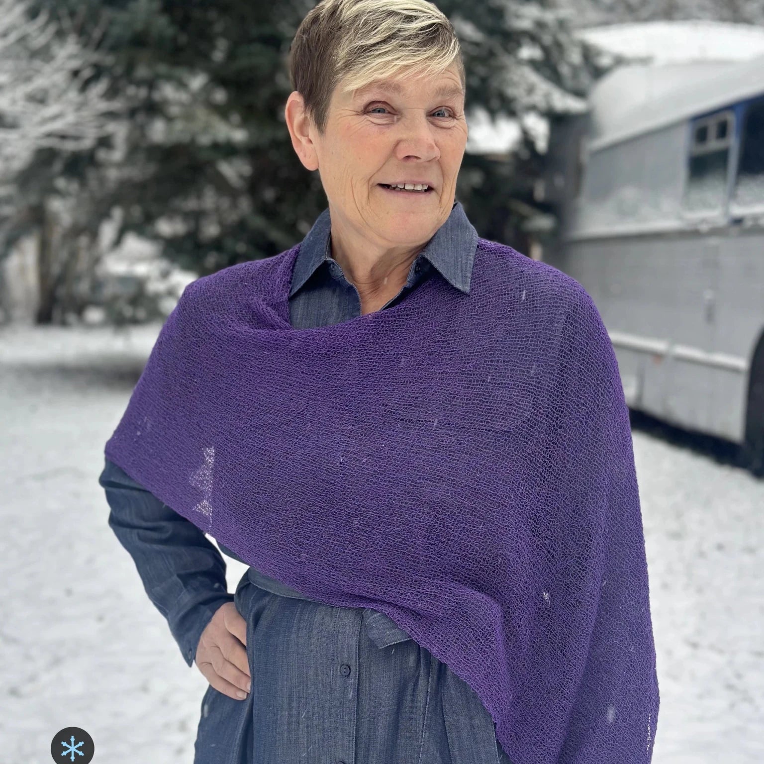 Kvinna bär Ella Cape i färg 065 Plommonlila, en kall färg, finstickad cape som draperas mjukt över axlarna, fotad en snöig dag. Ulla Jacobsson Kläder.