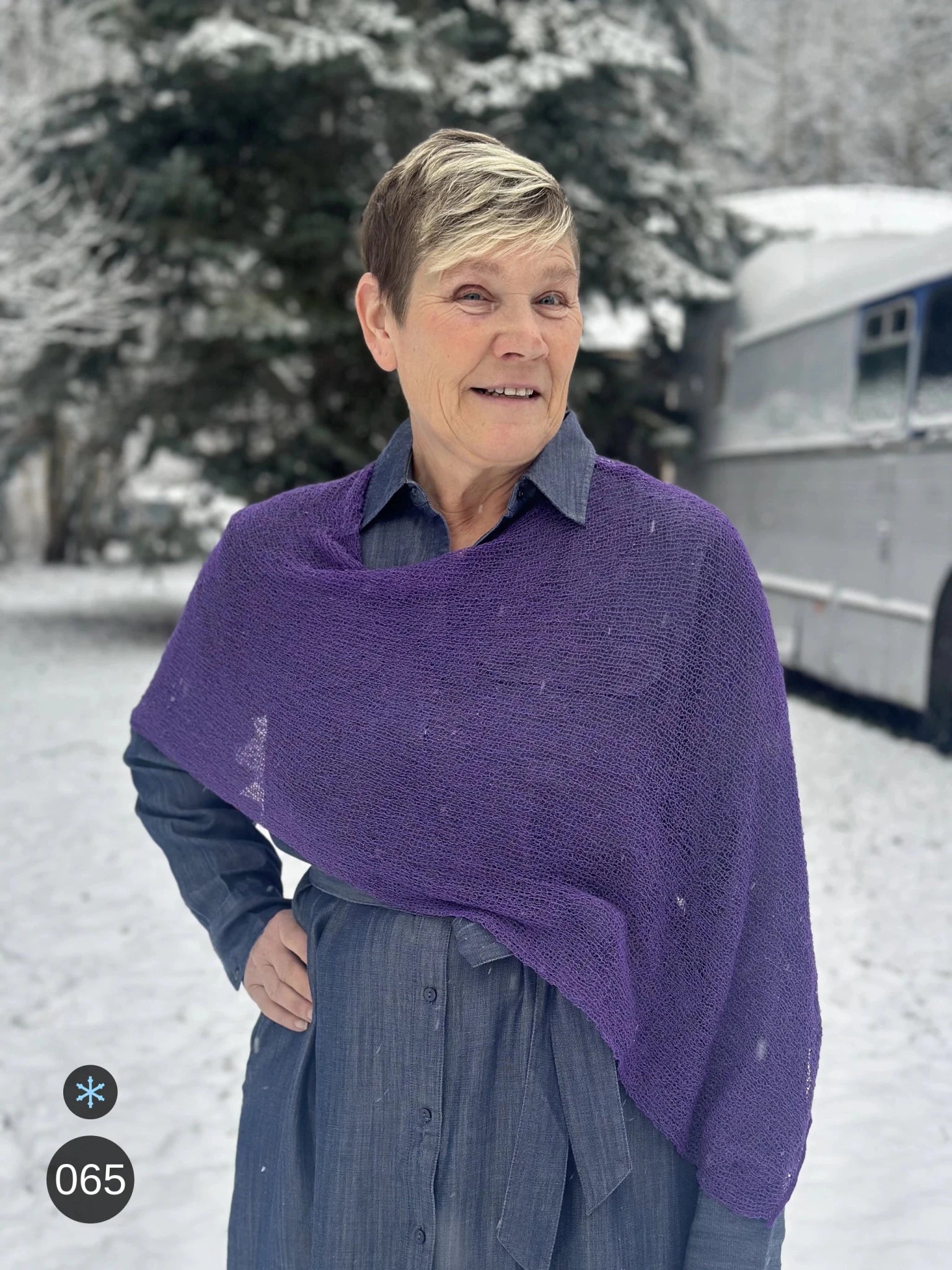 Kvinna bär Ella Cape i färg 065 Plommonlila, en kall färg, finstickad cape som draperas mjukt över axlarna, fotad en snöig dag. Ulla Jacobsson Kläder.