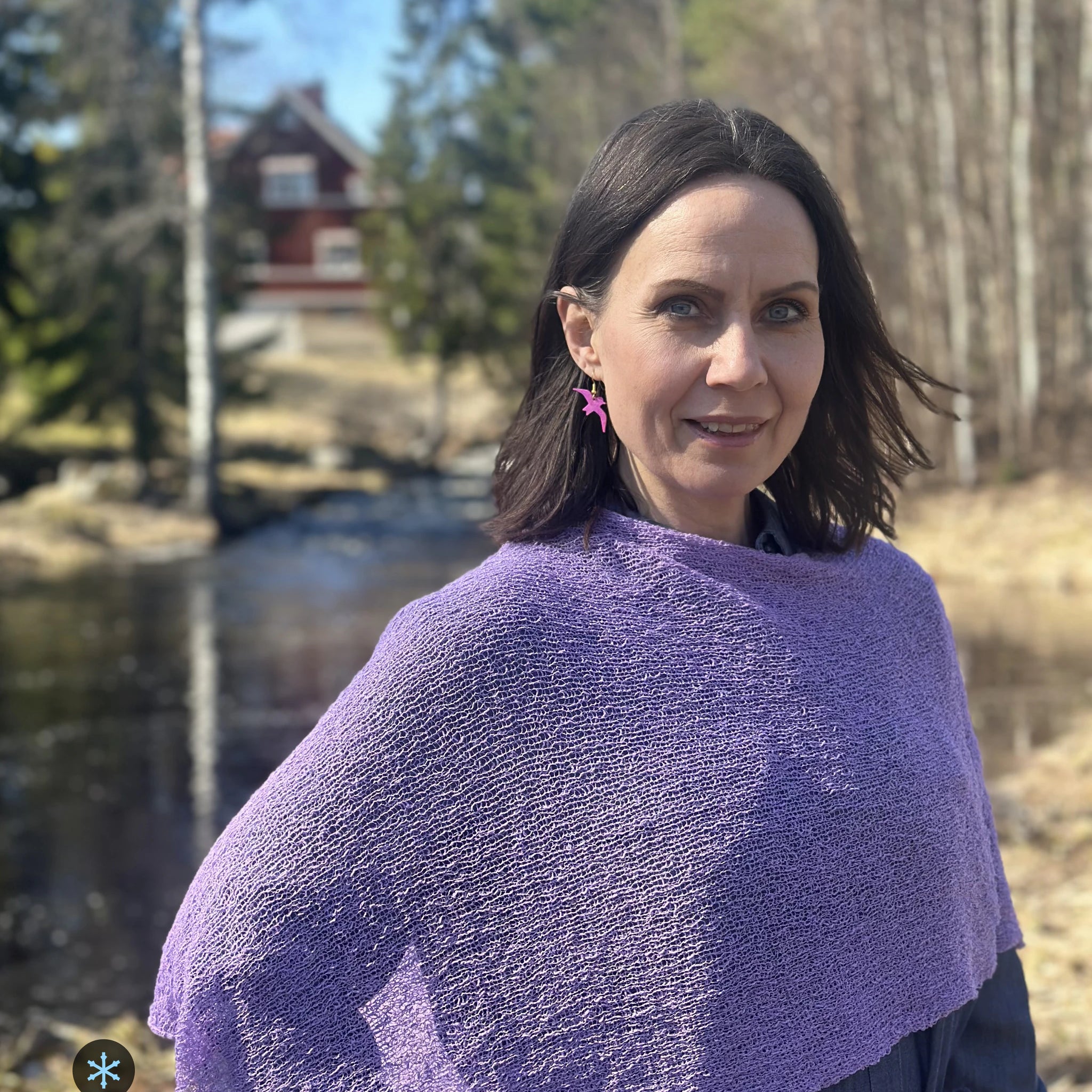 Kvinna bär Ella Cape i färg 067 Ljuslila, en kall färg, finstickad cape som draperas mjukt över axlarna, fotad vid en å i vårsol. Ulla Jacobsson Kläder.