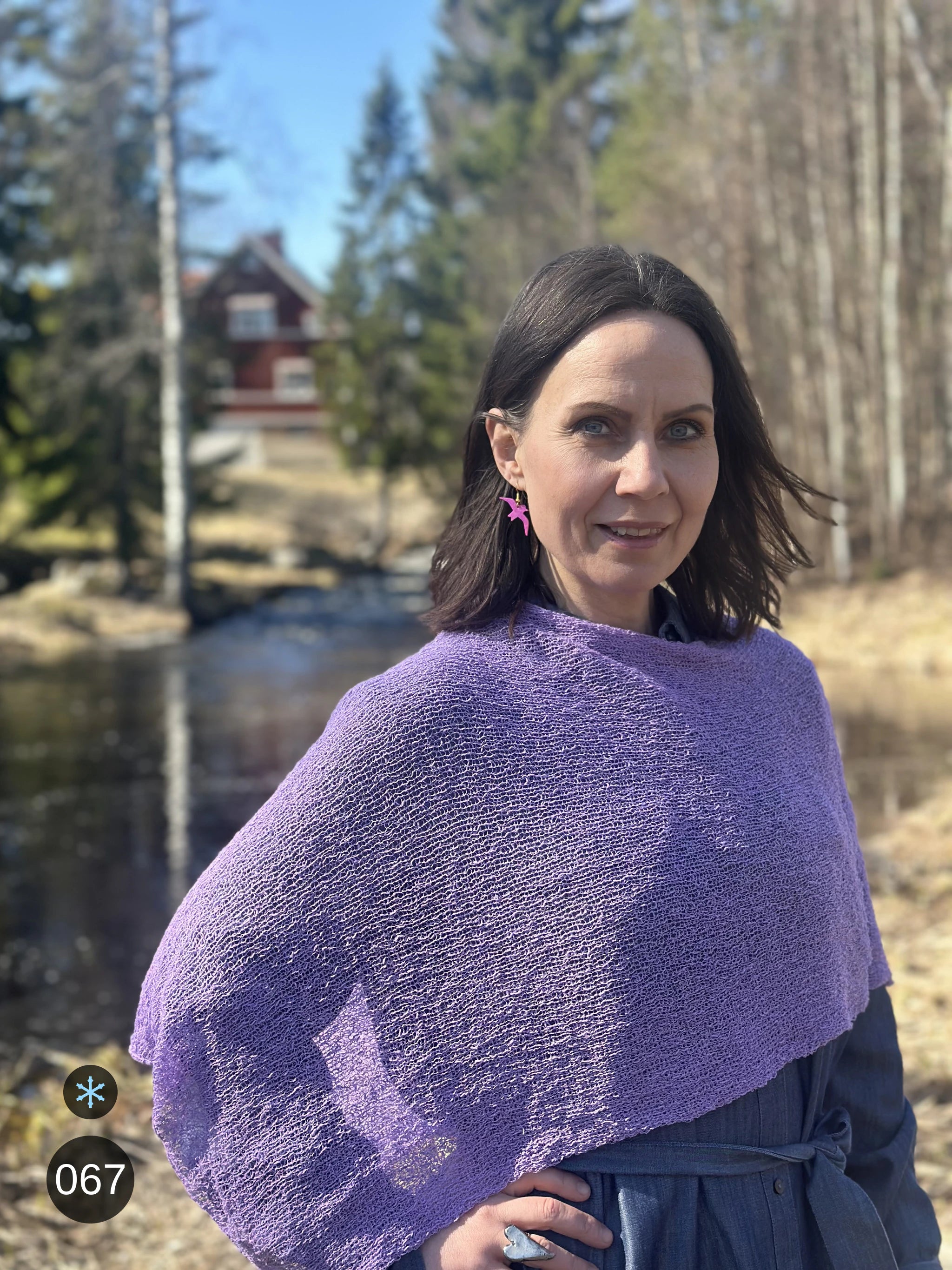 Kvinna bär Ella Cape i färg 067 Ljuslila, en kall färg, finstickad cape som draperas mjukt över axlarna, fotad vid en å i vårsol. Ulla Jacobsson Kläder.