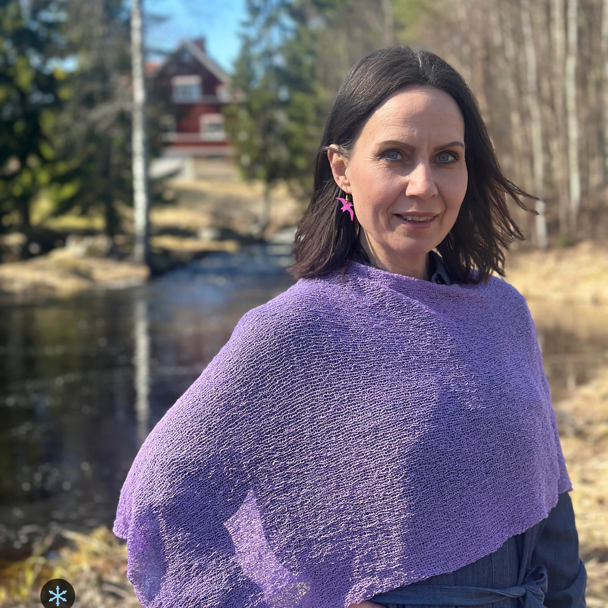 Kvinna bär Ella Cape i färg 067 Ljuslila, en kall färg i ljus, sval lila, finstickad cape som draperas mjukt över axlarna, fotad vid en å i vårsol. Ulla Jacobsson Kläder.