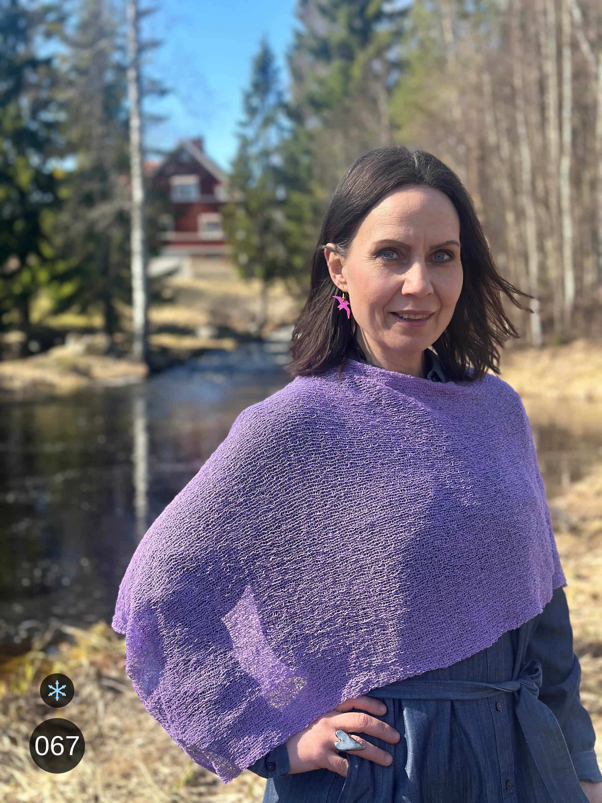 Kvinna bär Ella Cape i färg 067 Ljuslila, en kall färg i ljus, sval lila, finstickad cape som draperas mjukt över axlarna, fotad vid en å i vårsol. Ulla Jacobsson Kläder.
