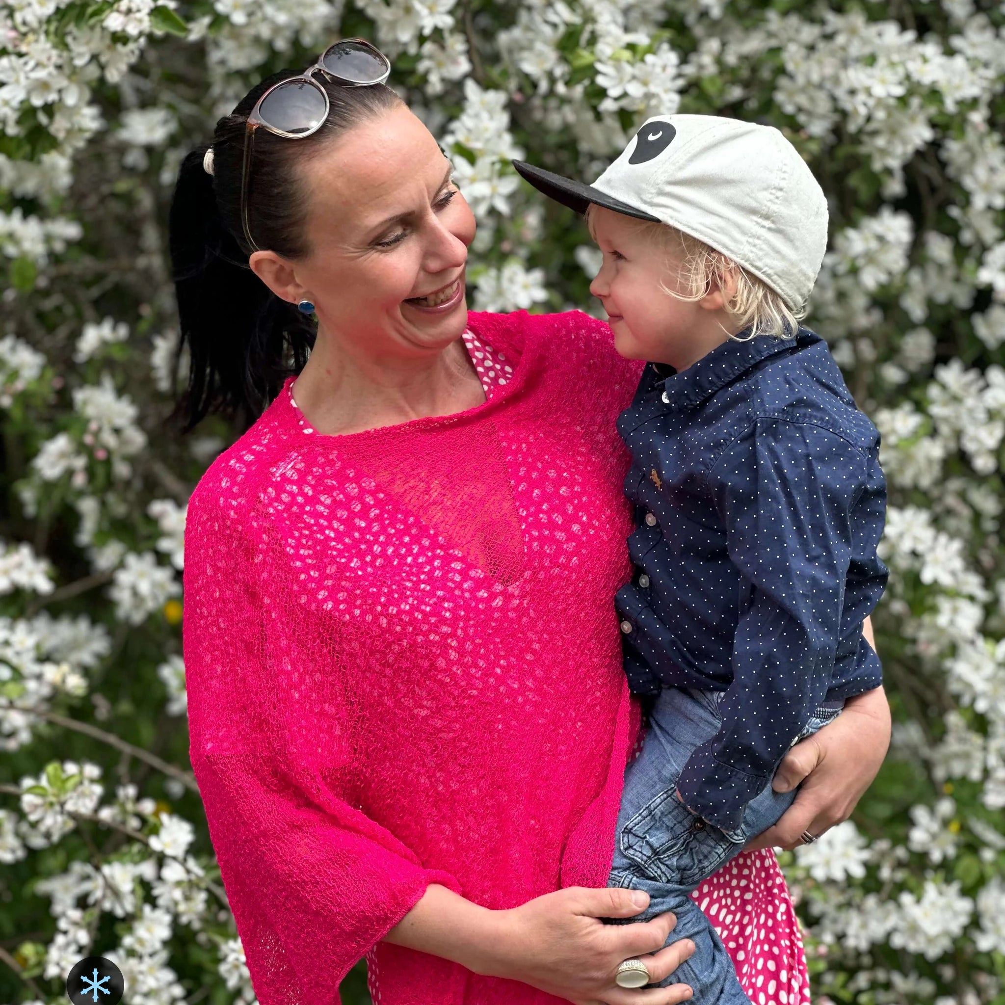 Kvinna bär Ella Cape i färg 070 Neonrosa, en kall färg i klar neonrosa, finstickad cape som draperas mjukt över axlarna, fotad bland vita vårblommor med ett barn i famnen. Ulla Jacobsson Kläder.