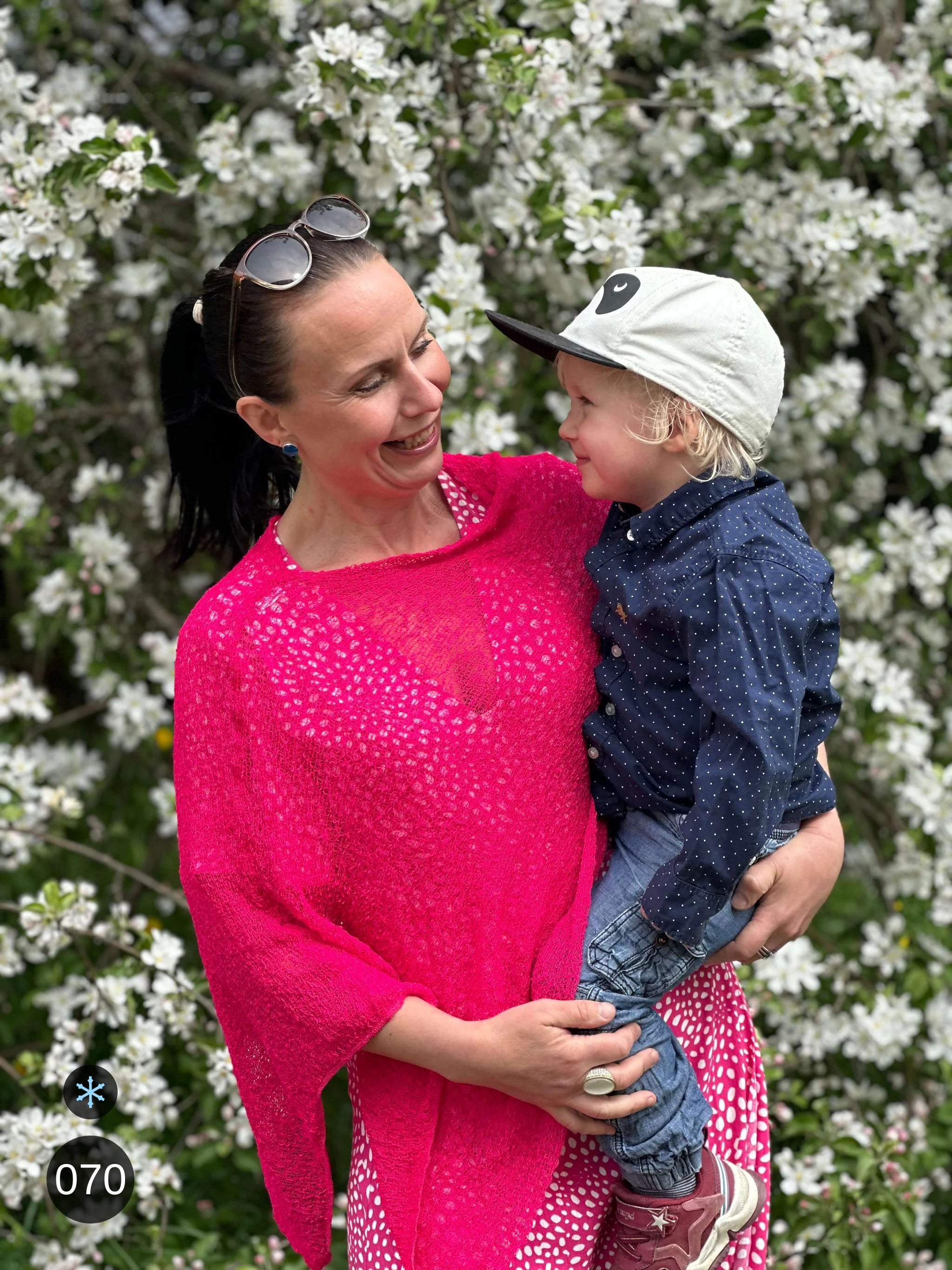 Kvinna bär Ella Cape i färg 070 Neonrosa, en kall färg i klar neonrosa, finstickad cape som draperas mjukt över axlarna, fotad bland vita vårblommor med ett barn i famnen. Ulla Jacobsson Kläder.