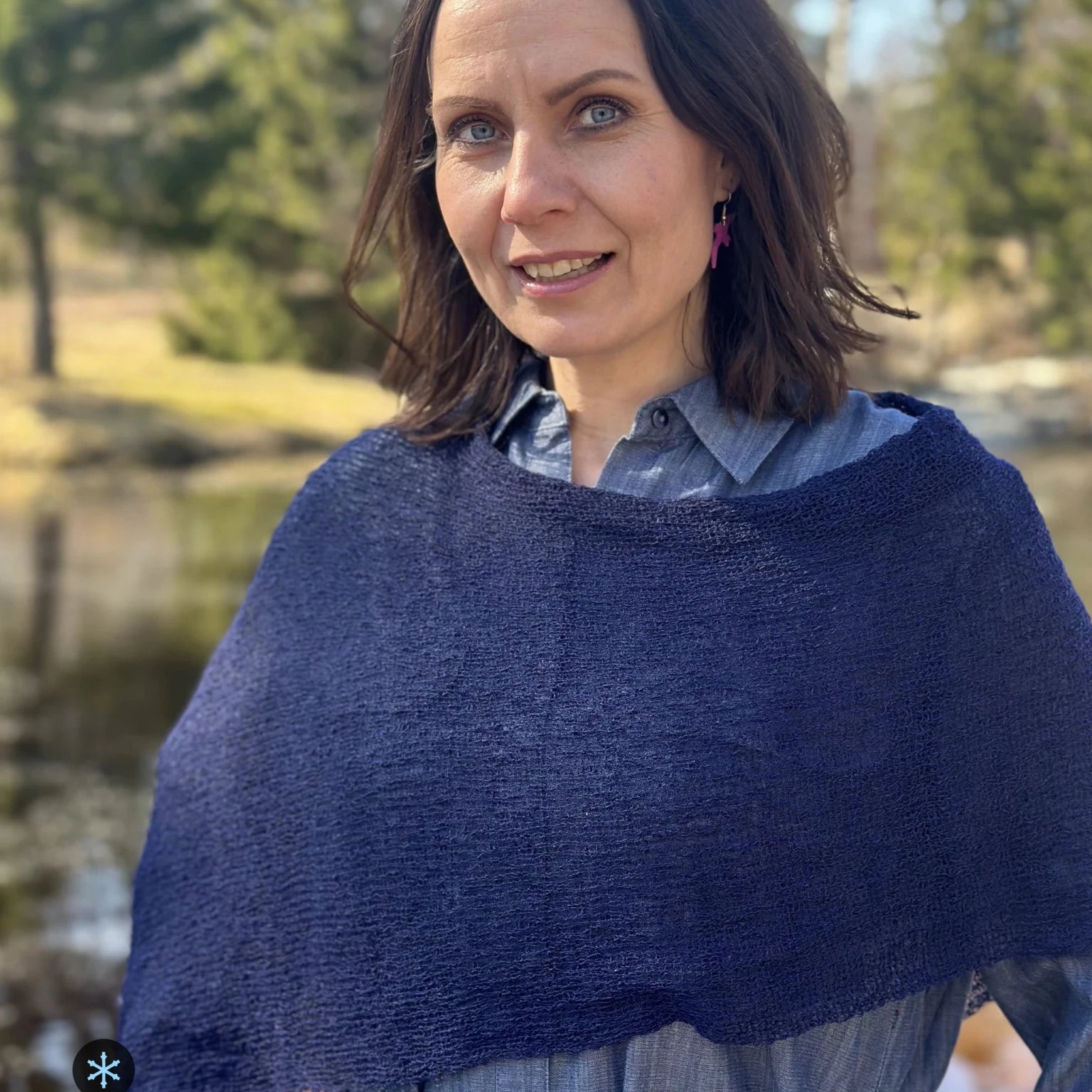 Kvinna bär Ella Cape i färg 107 Mörk marin, en kall färg, finstickad cape som draperas mjukt över axlarna, fotad utomhus vid vattnet i vårsol. Ulla Jacobsson Kläder.