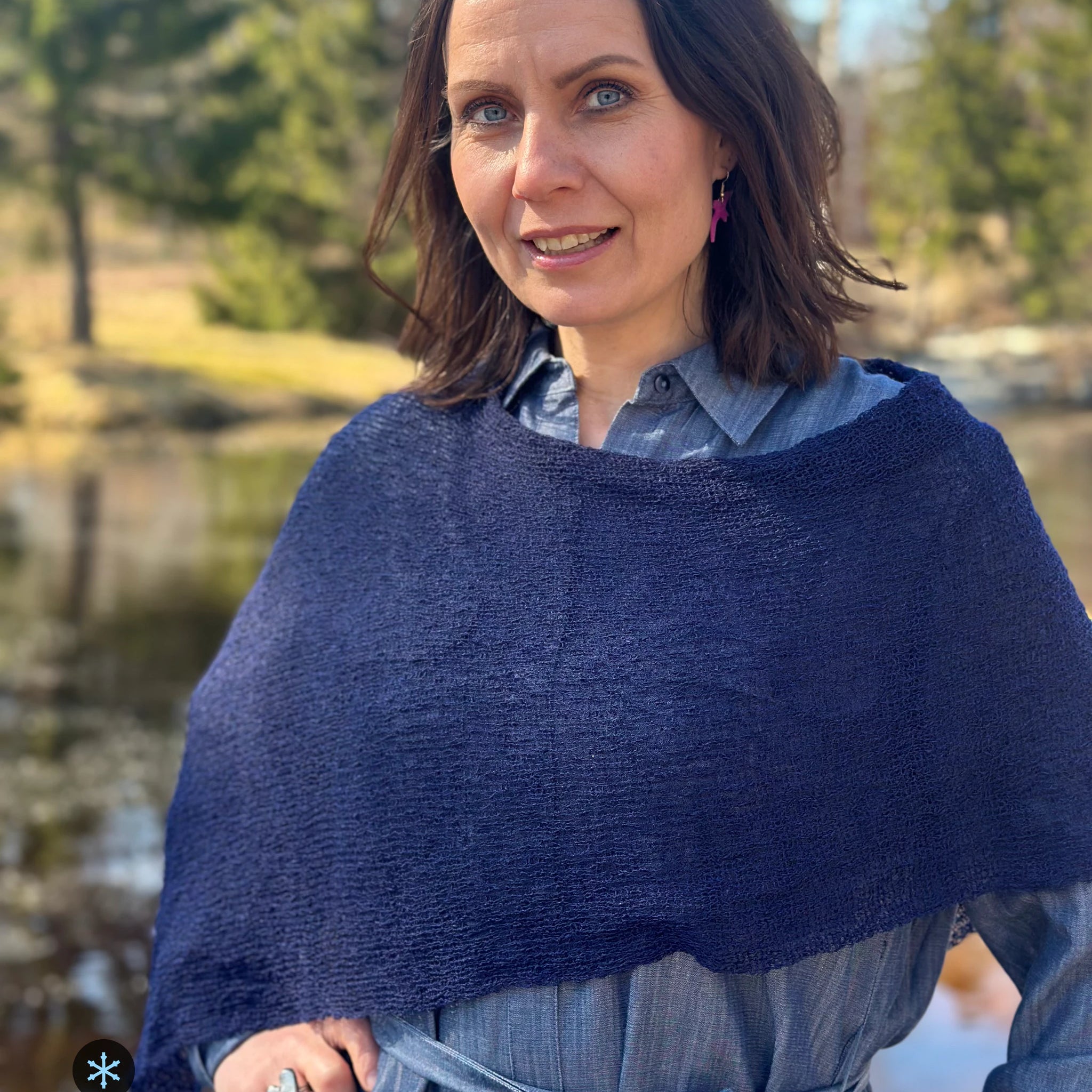 Kvinna bär Ella Cape i färg 107 Mörk marin, en kall färg i djup mörkblå marin, finstickad cape som draperas mjukt över axlarna, fotad vid en å en tidig vårdag. Ulla Jacobsson Kläder.