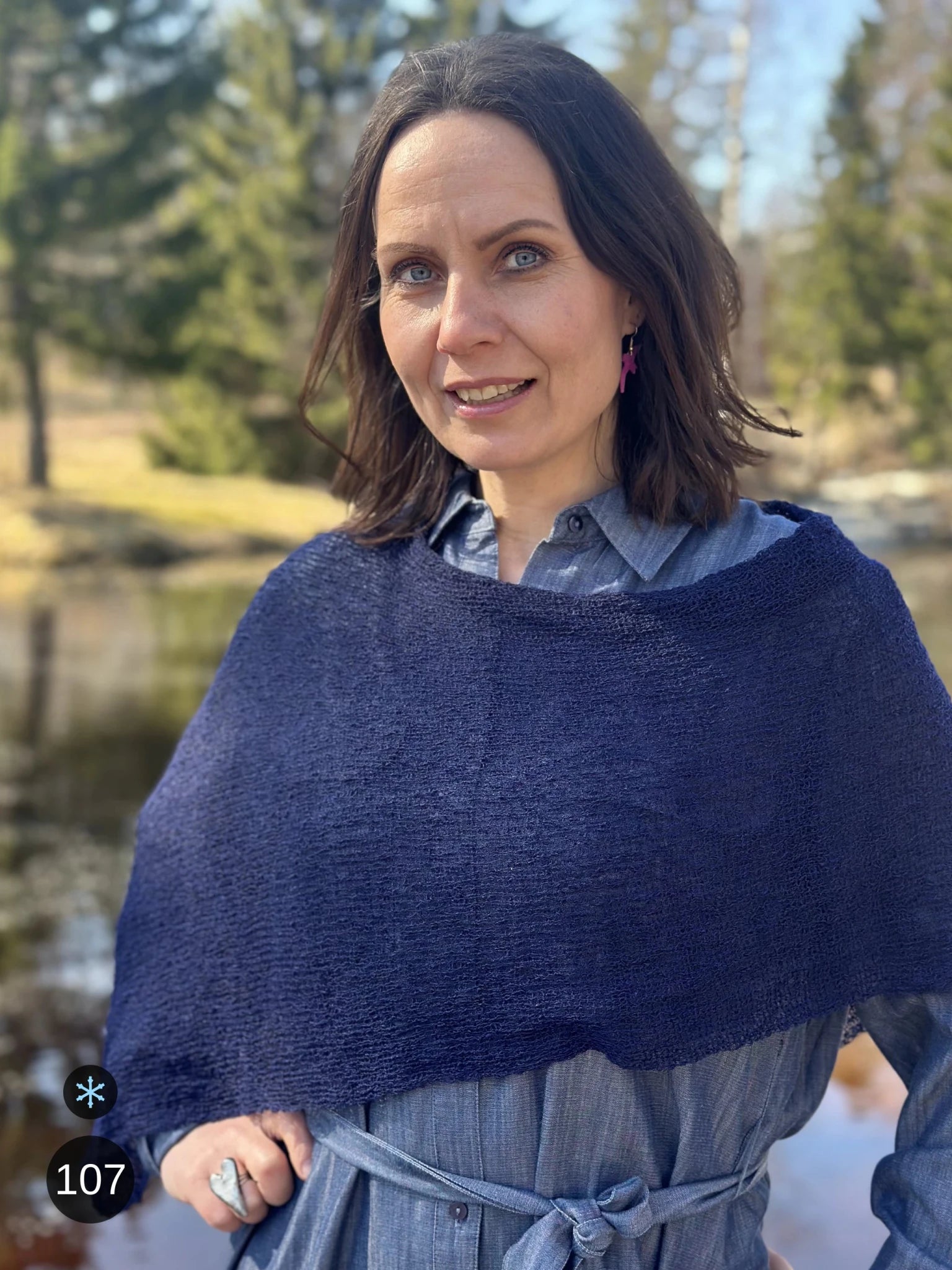 Kvinna bär Ella Cape i färg 107 Mörk marin, en kall färg, finstickad cape som draperas mjukt över axlarna, fotad utomhus vid vattnet i vårsol. Ulla Jacobsson Kläder.
