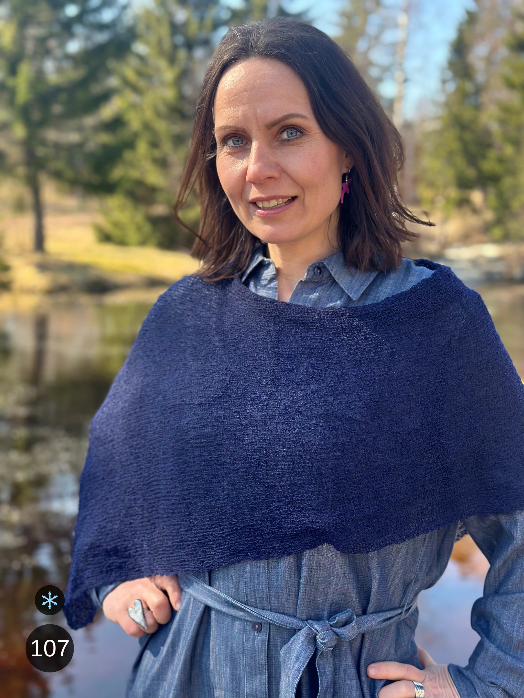 Kvinna bär Ella Cape i färg 107 Mörk marin, en kall färg i djup mörkblå marin, finstickad cape som draperas mjukt över axlarna, fotad vid en å en tidig vårdag. Ulla Jacobsson Kläder.