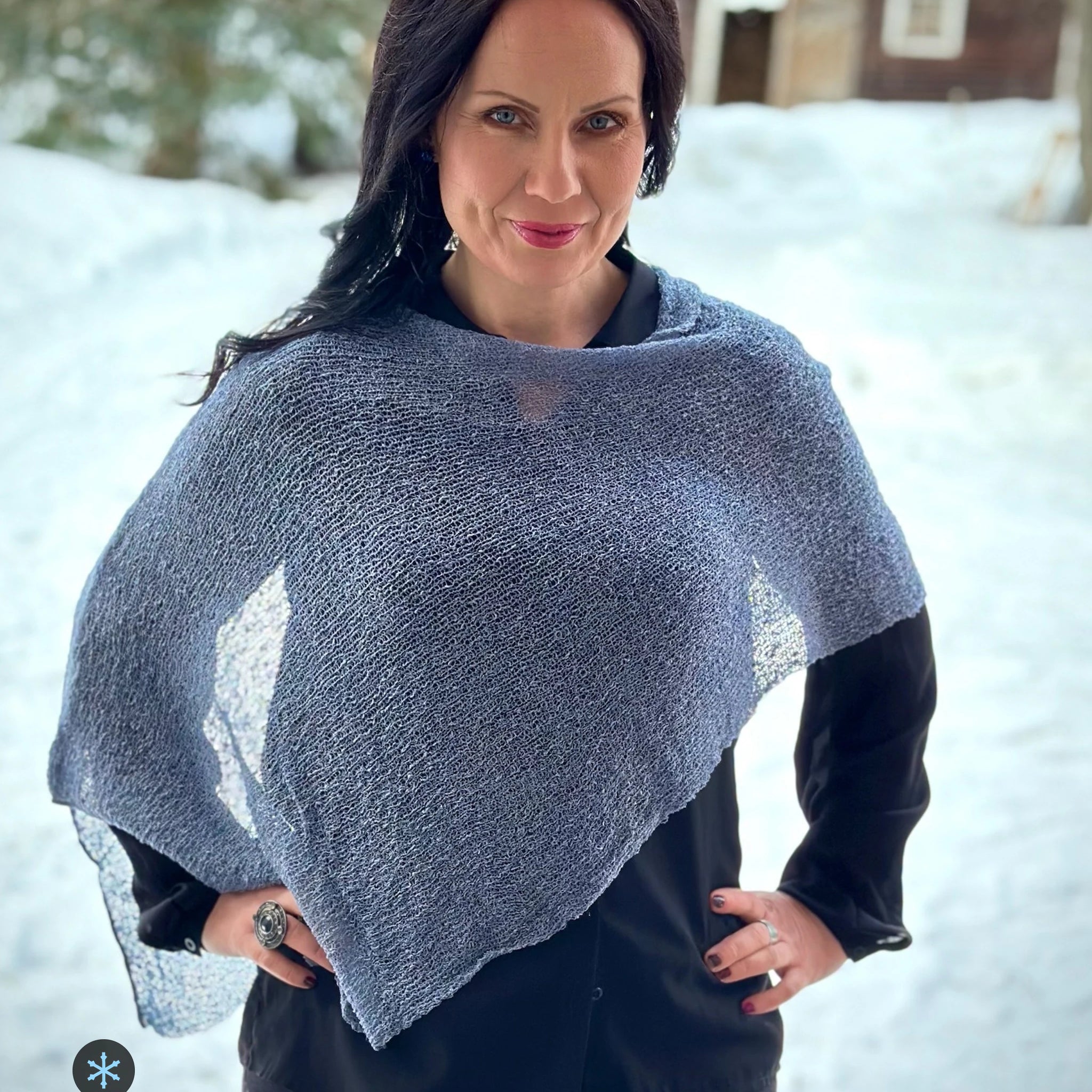 Kvinna bär Ella Cape i färg 108 Granit, en kall färg i granitgrå med sval blå underton, finstickad cape som draperas mjukt över axlarna, fotad mot snön framför en röd byggnad. Ulla Jacobsson Kläder.